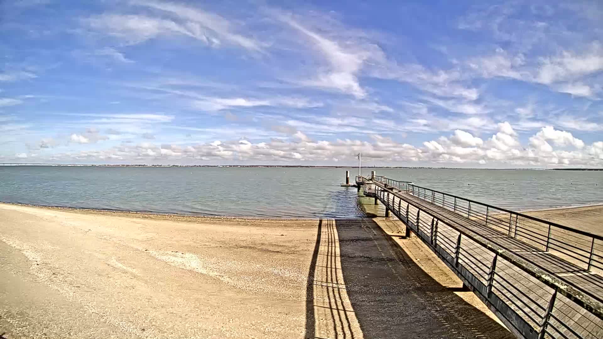 A sandy beach stretches to a calm body of water under a mostly sunny sky with scattered clouds, where a long pier extends from the shore.