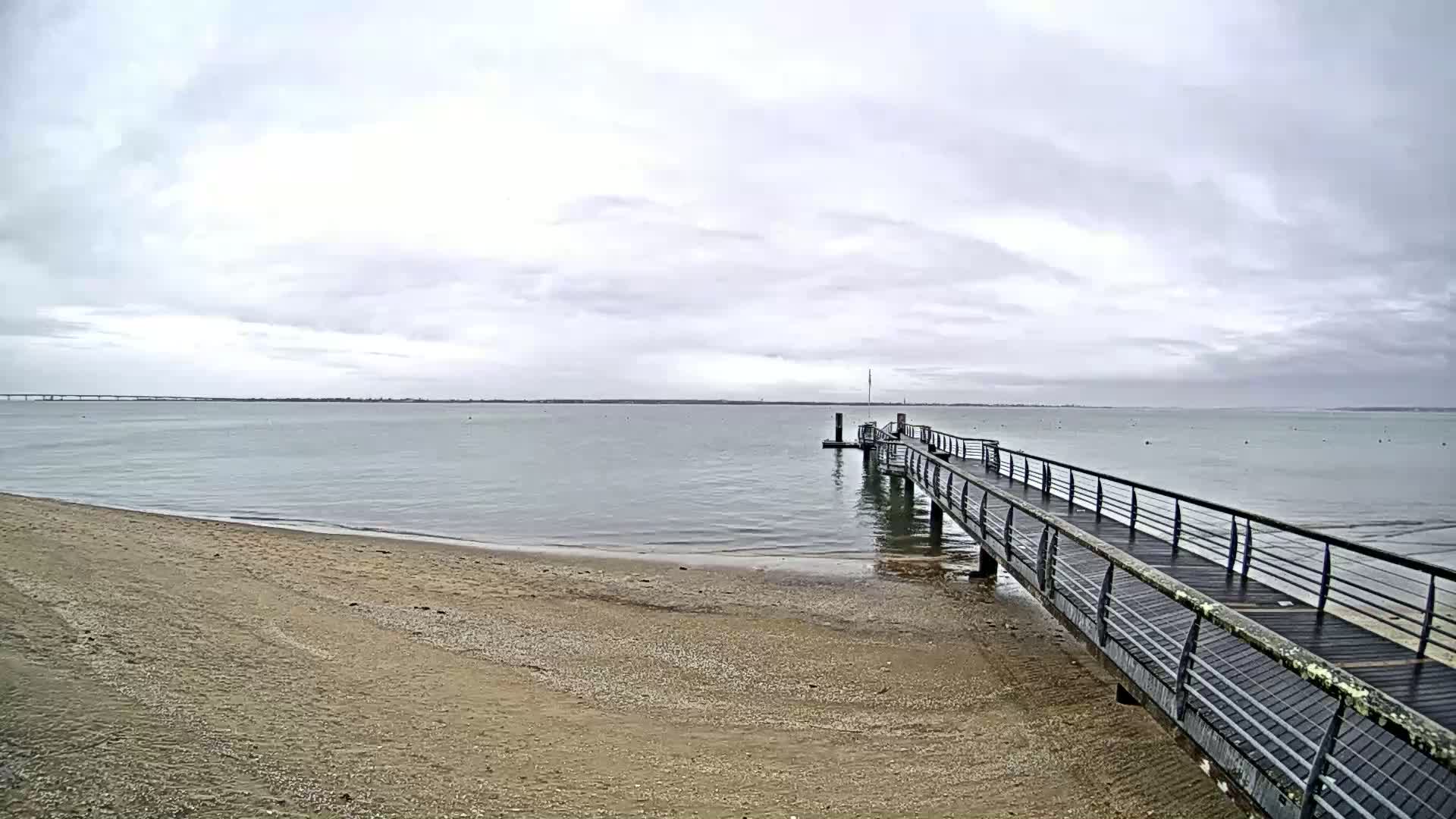 A long pier extends from a sandy beach into calm, gray water under a cloudy sky.