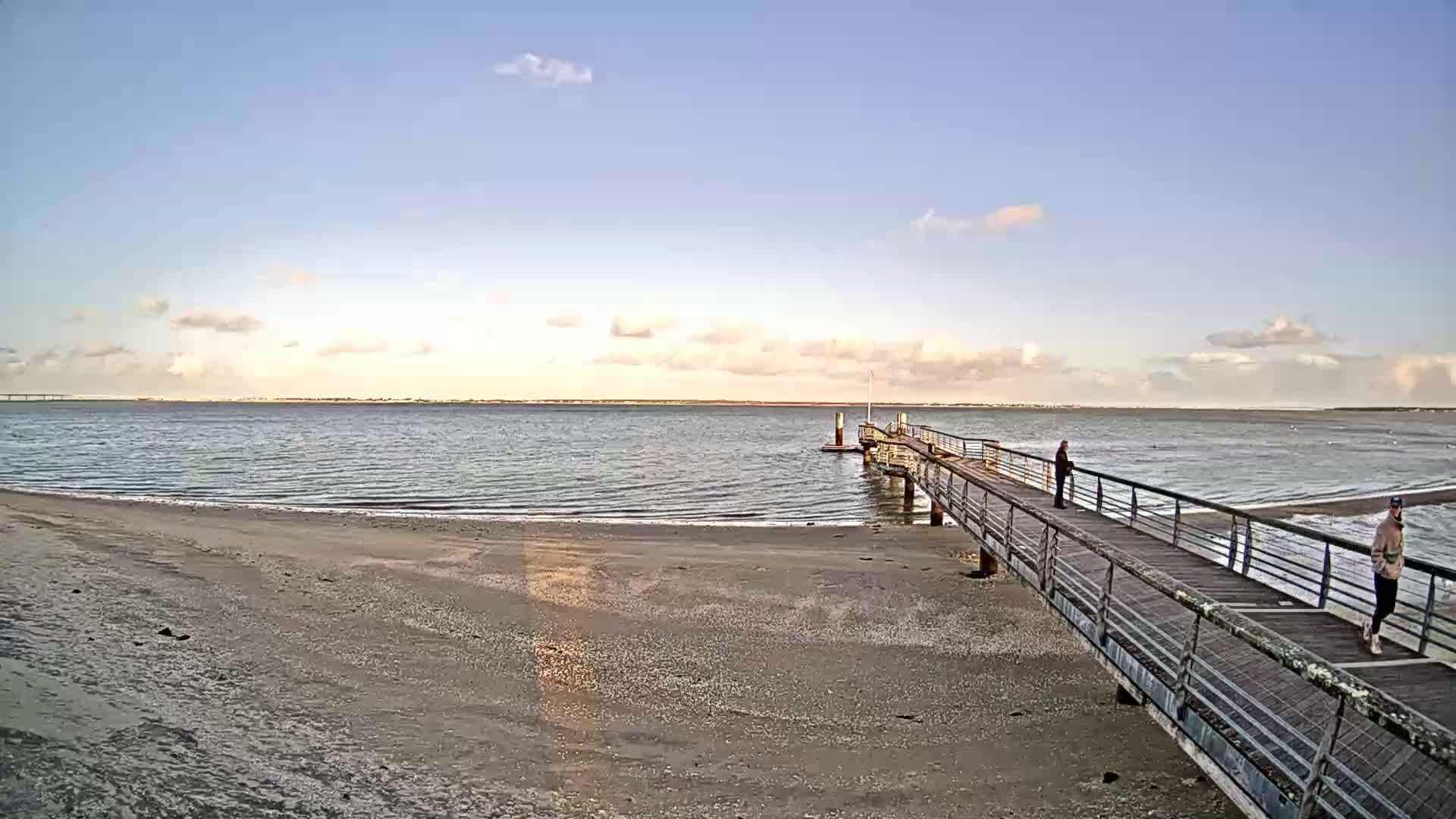 A long pier extends from a sandy beach into calm, light-blue water under a partly cloudy sky.
