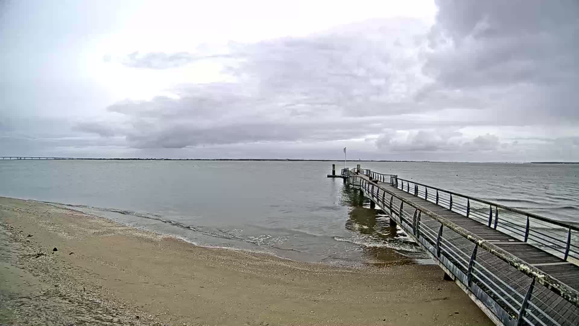A long pier extends from a sandy beach into a calm, gray body of water under a cloudy sky.