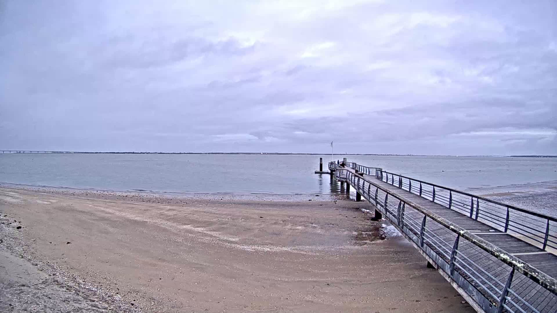 A long pier extends from a sandy beach into a calm body of water under an overcast sky.