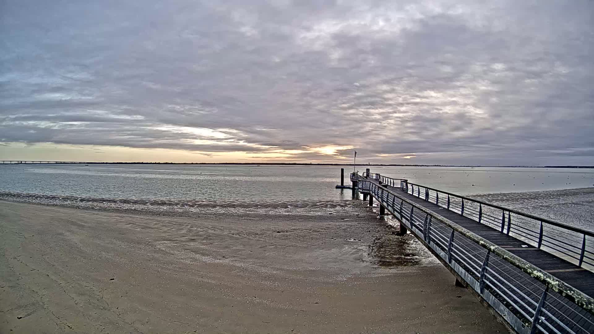 A long pier extends from a sandy beach into a calm body of water under a cloudy sky with a hint of sunset.