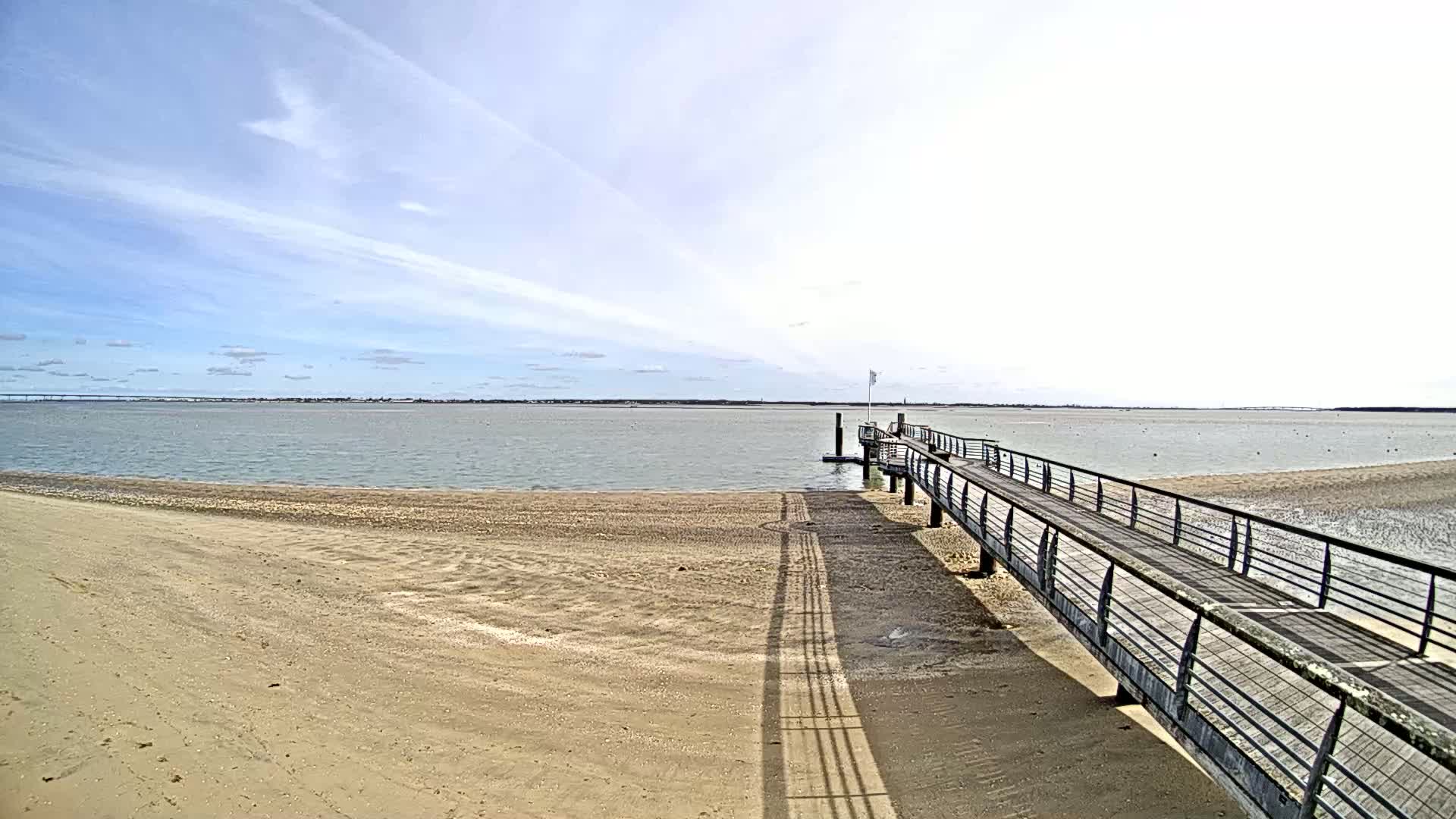 A long wooden pier extends from a sandy beach into a calm, light-grey body of water under a mostly clear, bright sky.