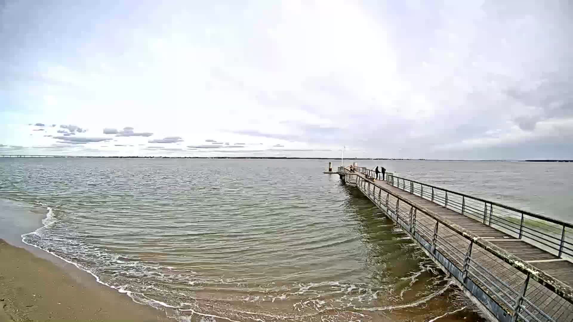 A long pier extends into a calm, grey sea under an overcast sky, with a few people standing near its end and small waves lapping at a sandy shore.