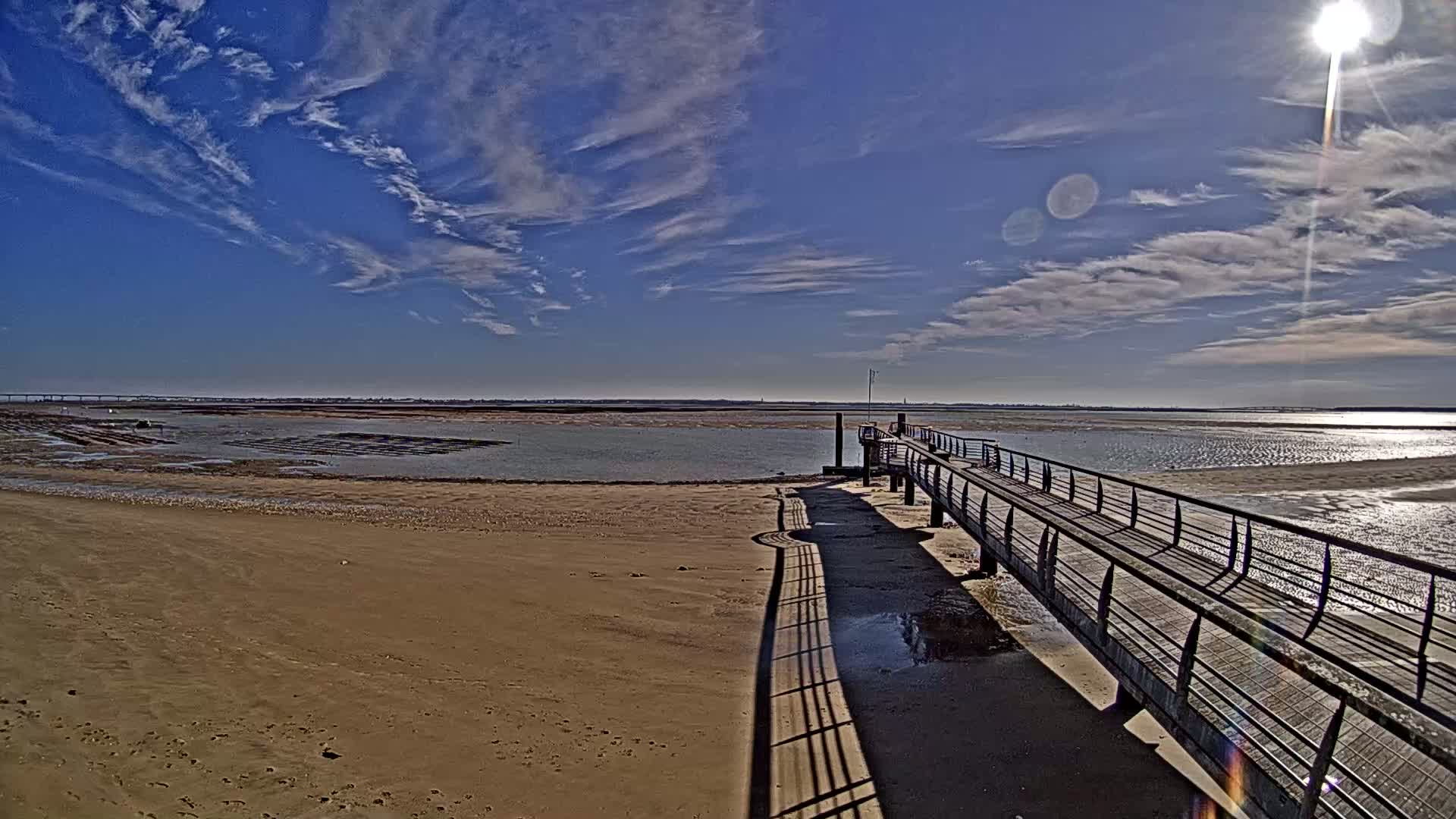 A long pier extends from a sandy beach into a calm body of water under a bright, sunny sky with scattered clouds.