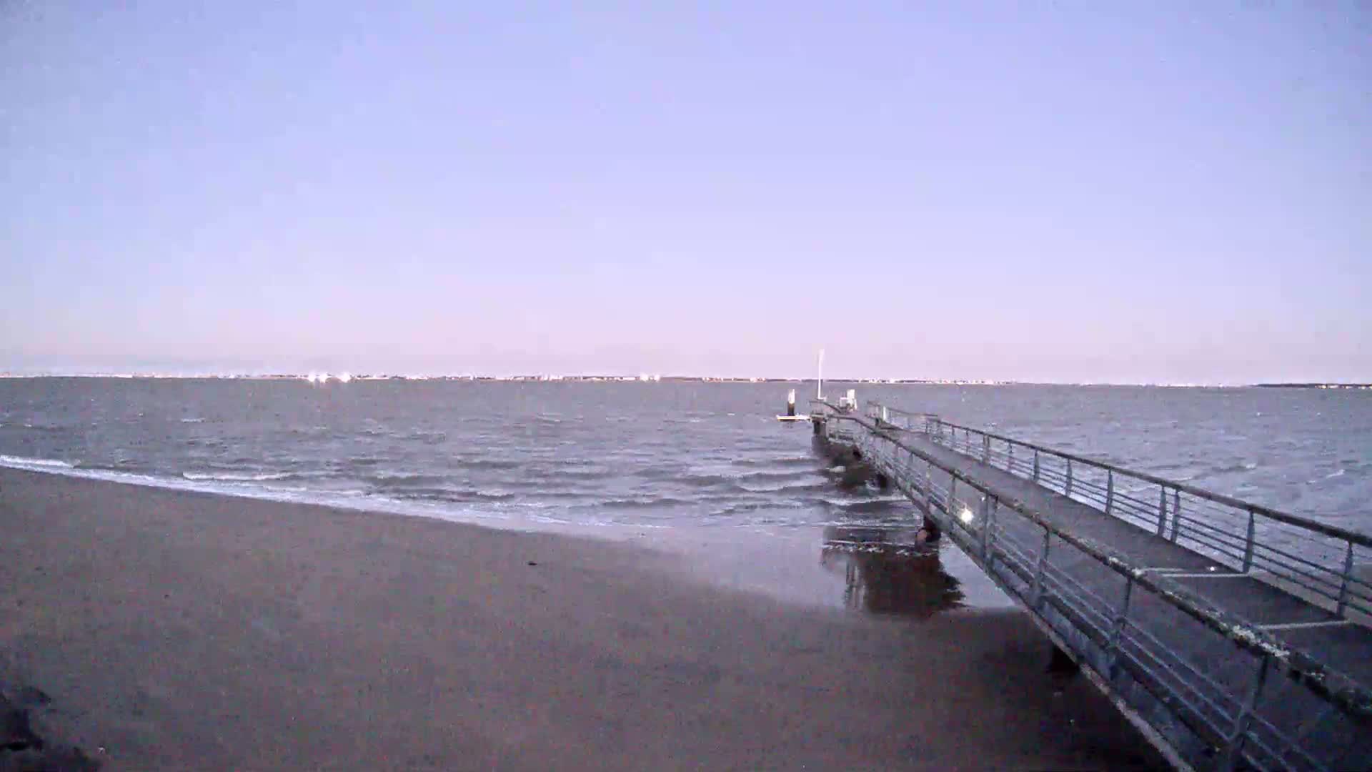 A long pier extends from a sandy beach into a calm, gray-blue sea under a pale, clear sky.