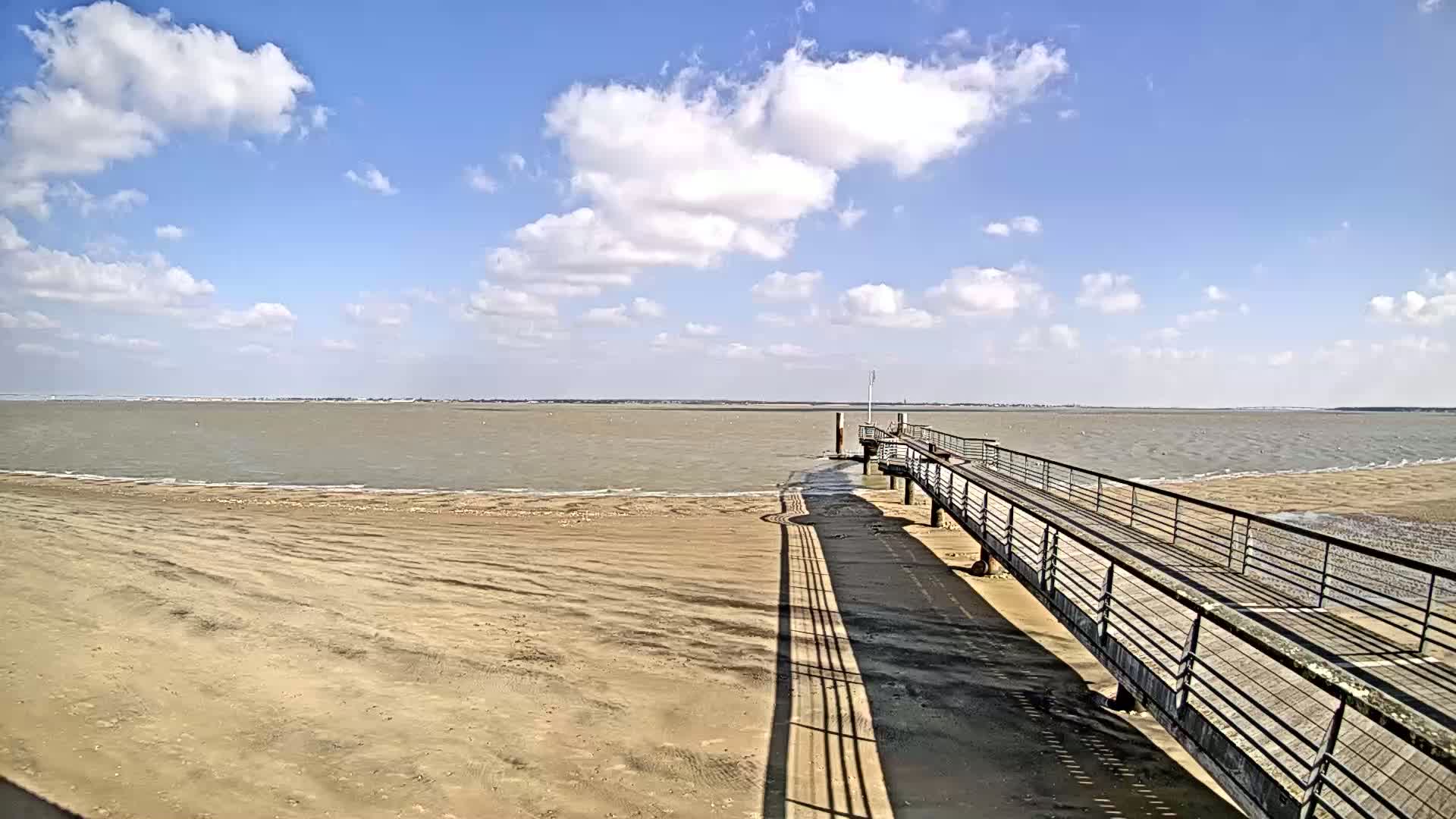 A long pier extends from a sandy beach into a calm, light-brown body of water under a mostly sunny sky with scattered cumulus clouds.
