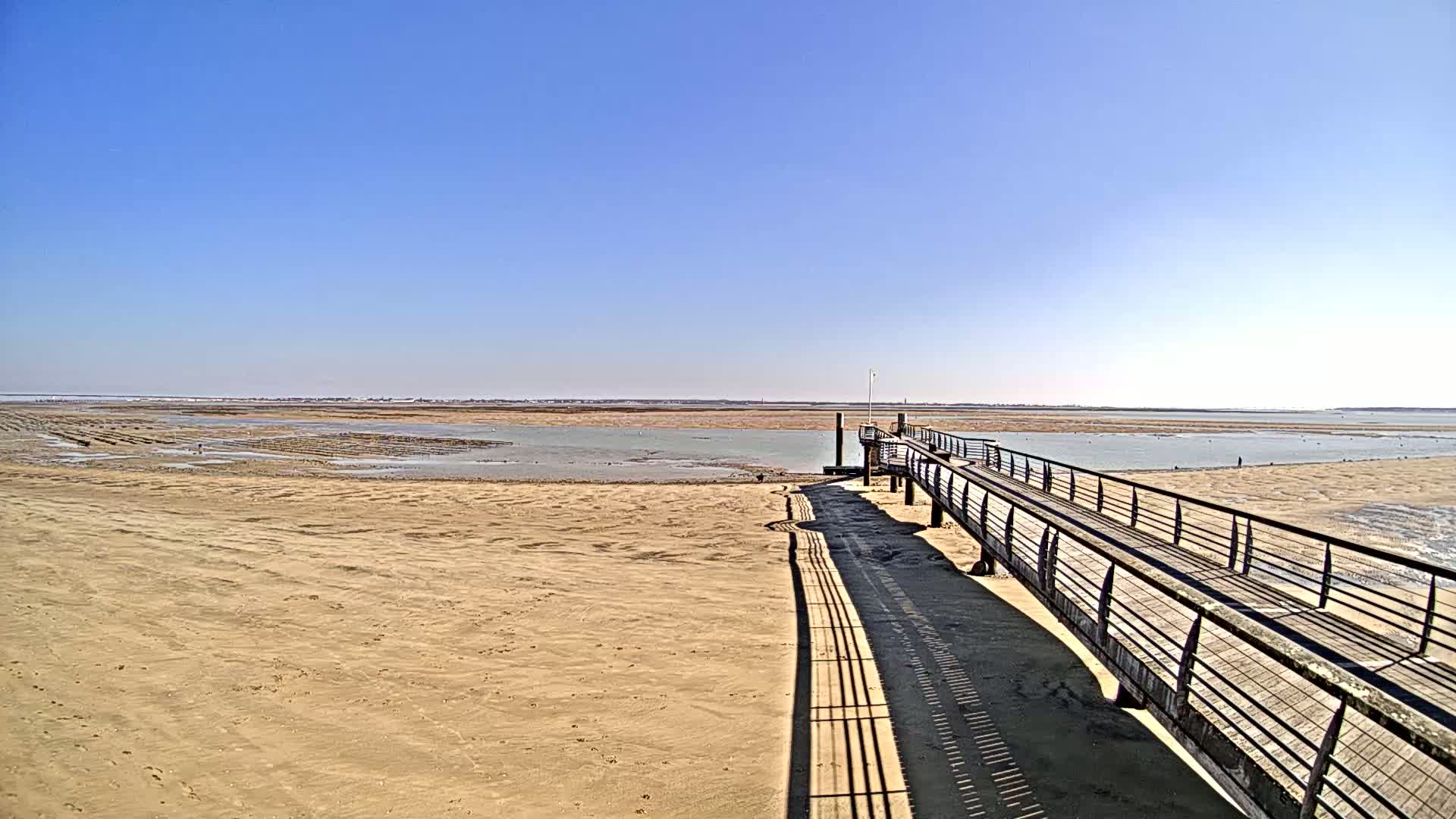 A long, narrow wooden pier stretches out over a sandy expanse towards a calm body of water under a clear blue sky.