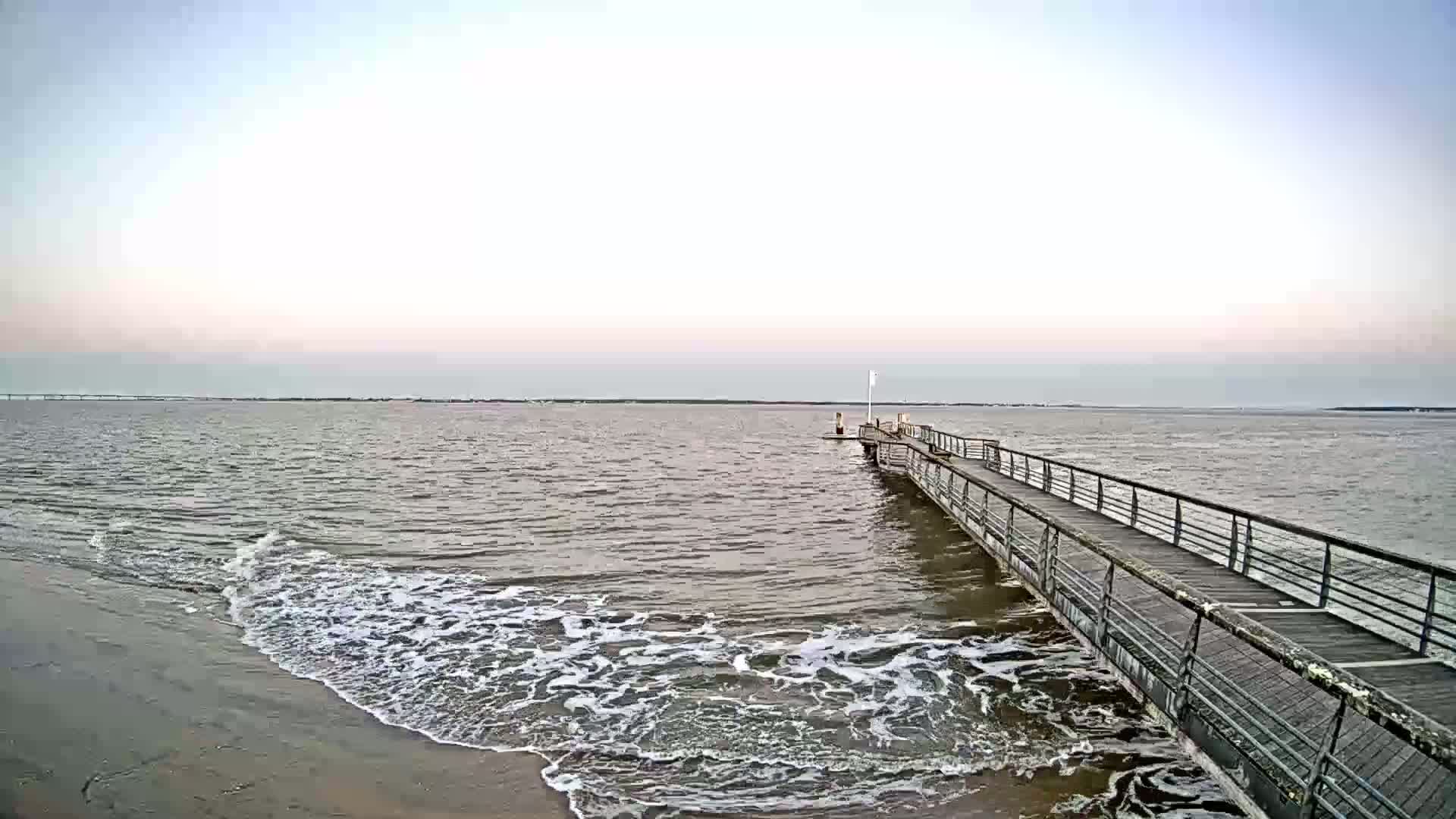 A long pier extends into calm, gray water with small waves lapping at a sandy shore under a pale sky.
