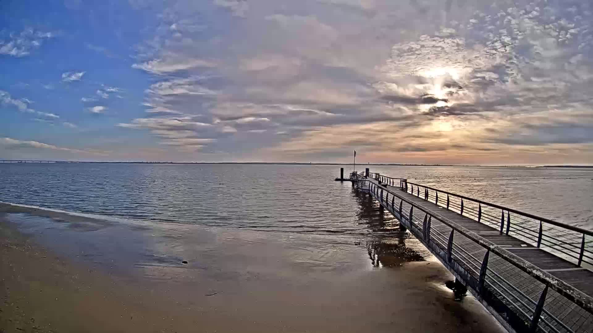 A long pier extends into a calm body of water under a partly cloudy sky with a bright sun shining through the clouds.