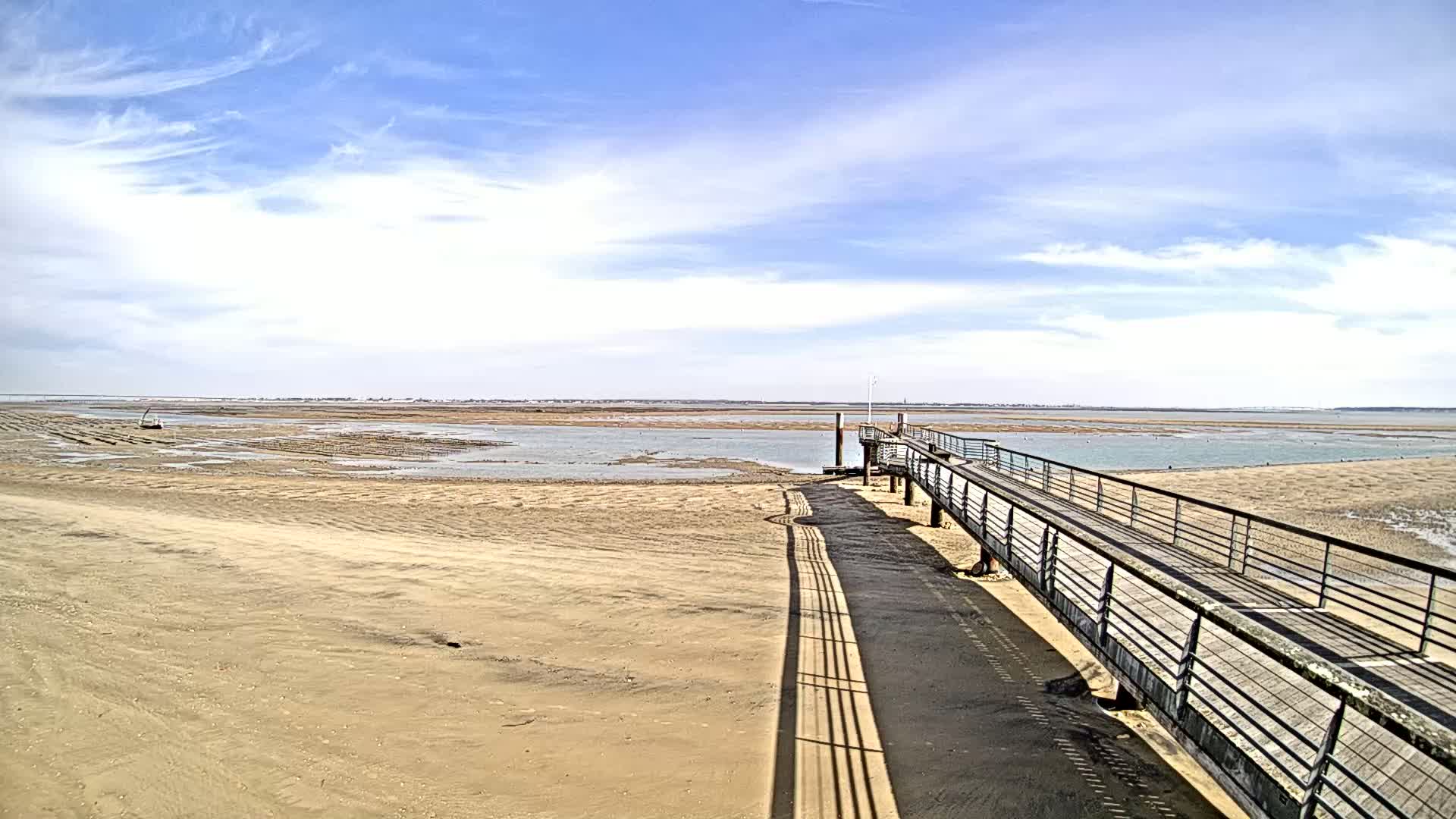 A long boardwalk extends from a sandy beach toward a calm body of water under a mostly sunny sky with scattered clouds.