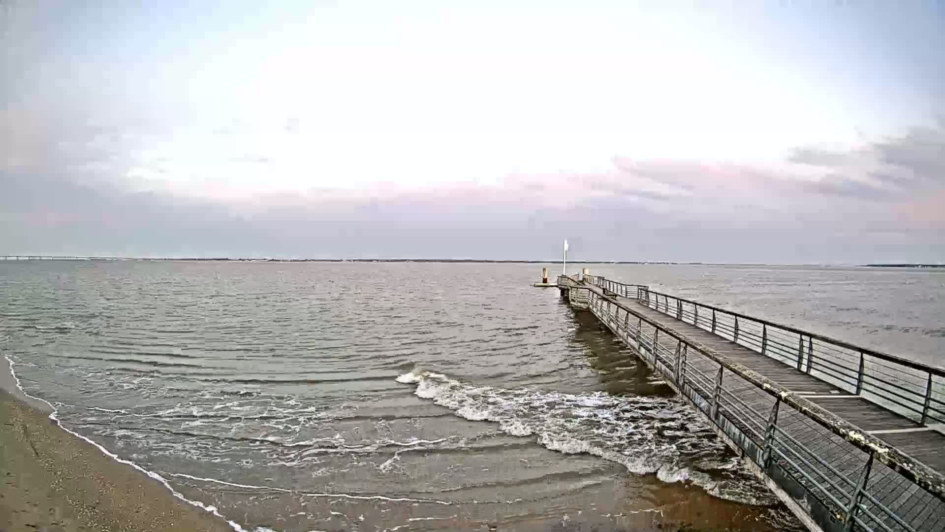 A long pier extends into a calm, gray body of water under a cloudy sky, with small waves lapping at a sandy shore.