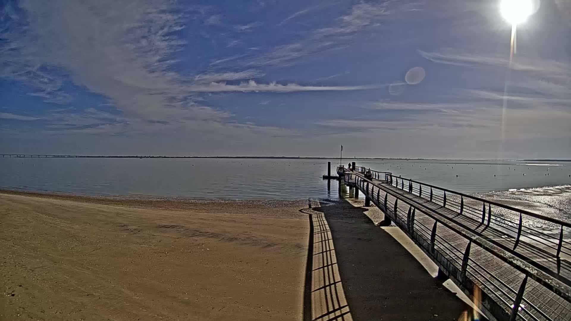 A sandy beach stretches to a calm body of water under a partly cloudy, sunny sky, with a long pier extending from the shore.