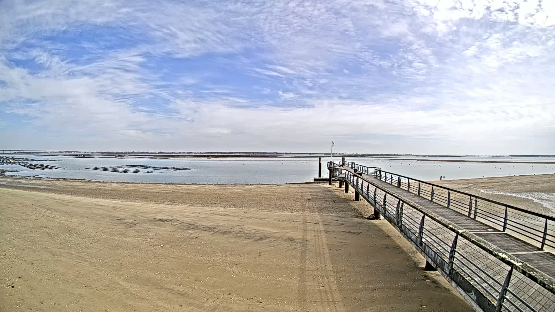 A long pier extends from a sandy beach into calm, shallow water under a mostly sunny sky with scattered clouds, overlooking oyster beds in the distance.