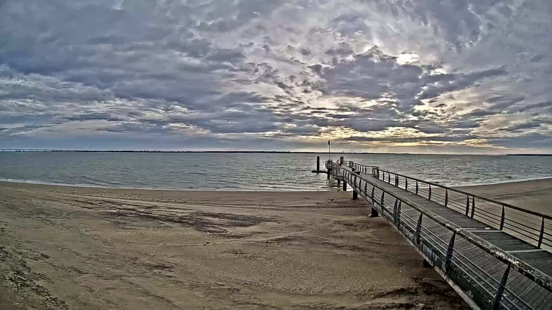 A long pier extends from a sandy beach into a calm body of water under a cloudy sky with hints of sunrise.
