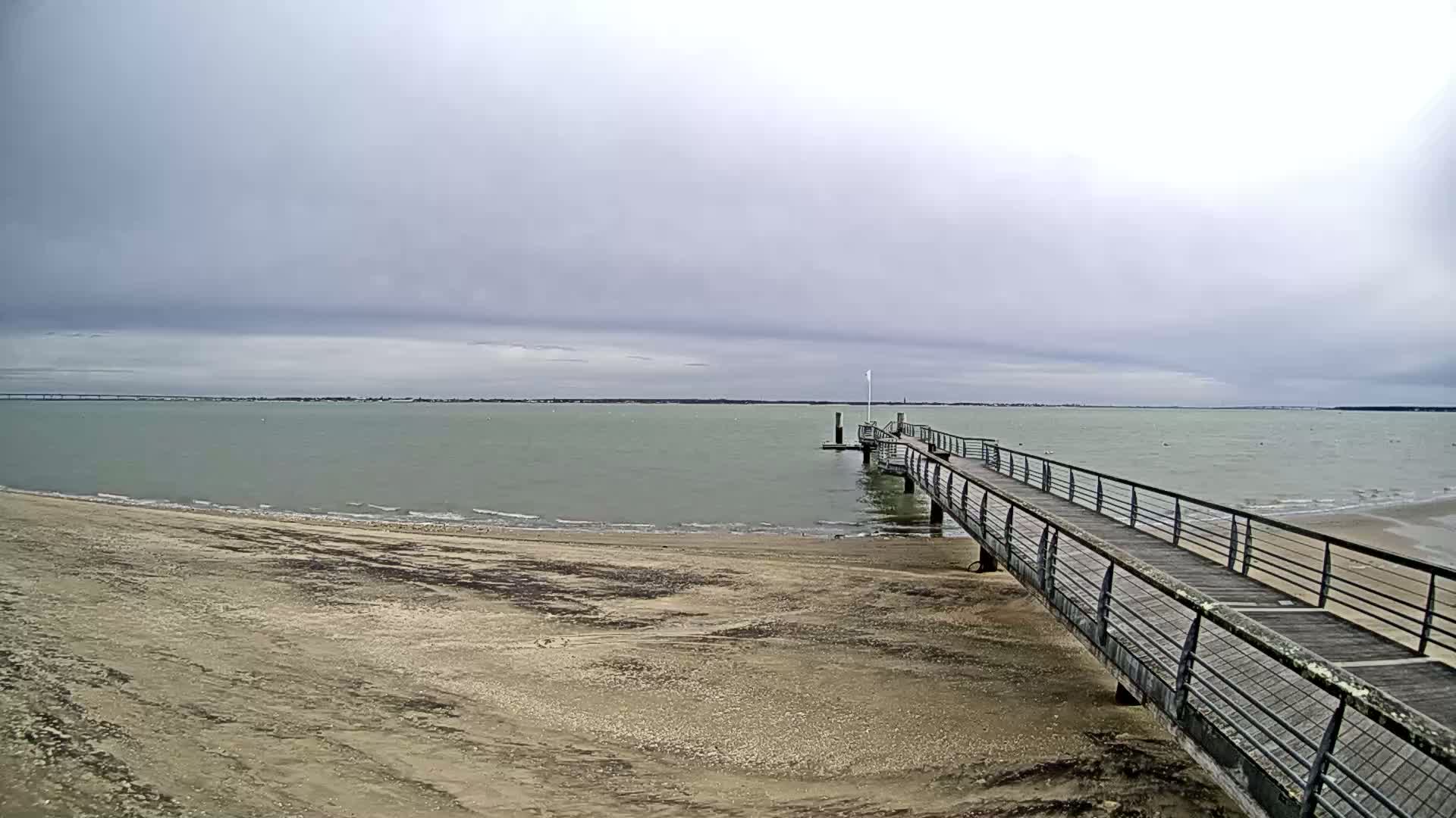 A long pier extends from a sandy beach into a calm, light-green body of water under an overcast sky.