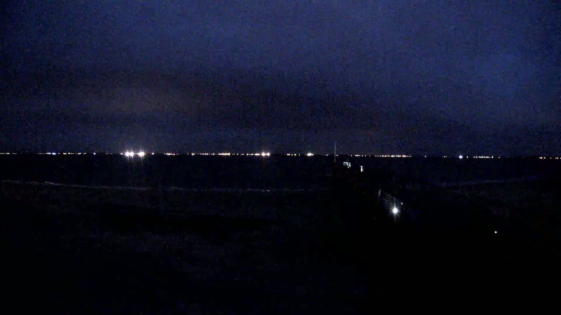 A dark, nighttime scene shows a body of water with a distant line of lights on the horizon under a dark, cloudy sky.