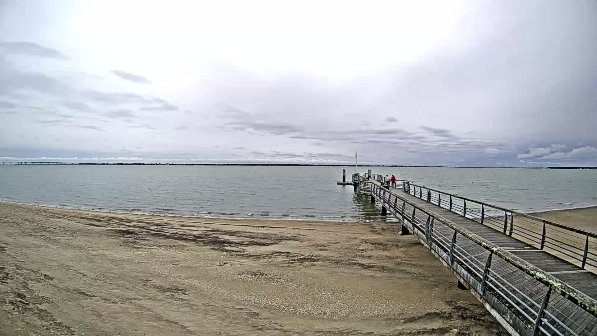 A long pier extends from a sandy beach into a calm, gray-cloudy day ocean, with two people walking towards the end of it.