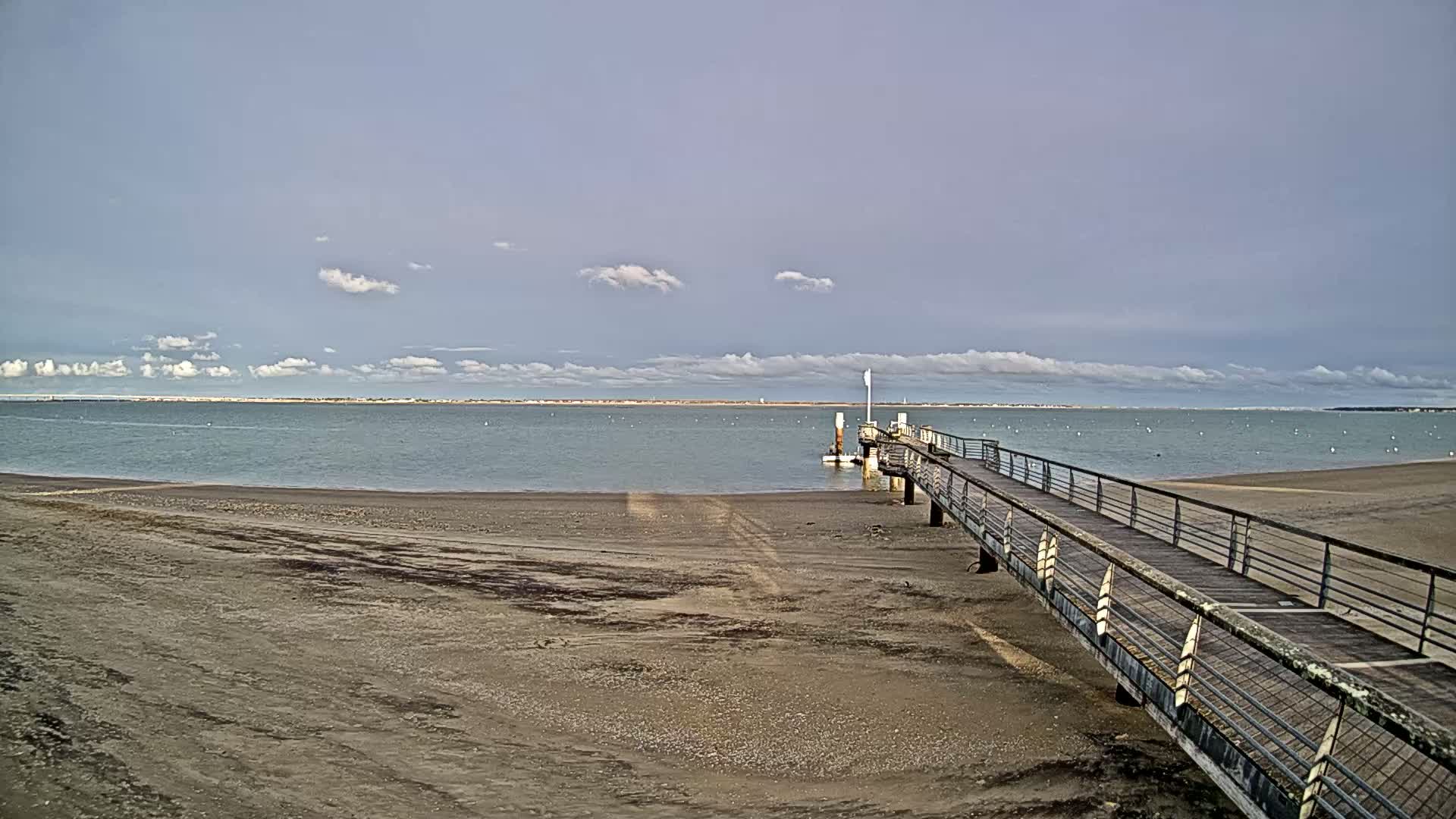 A long pier extends from a sandy beach into a calm body of water under a mostly cloudy sky.
