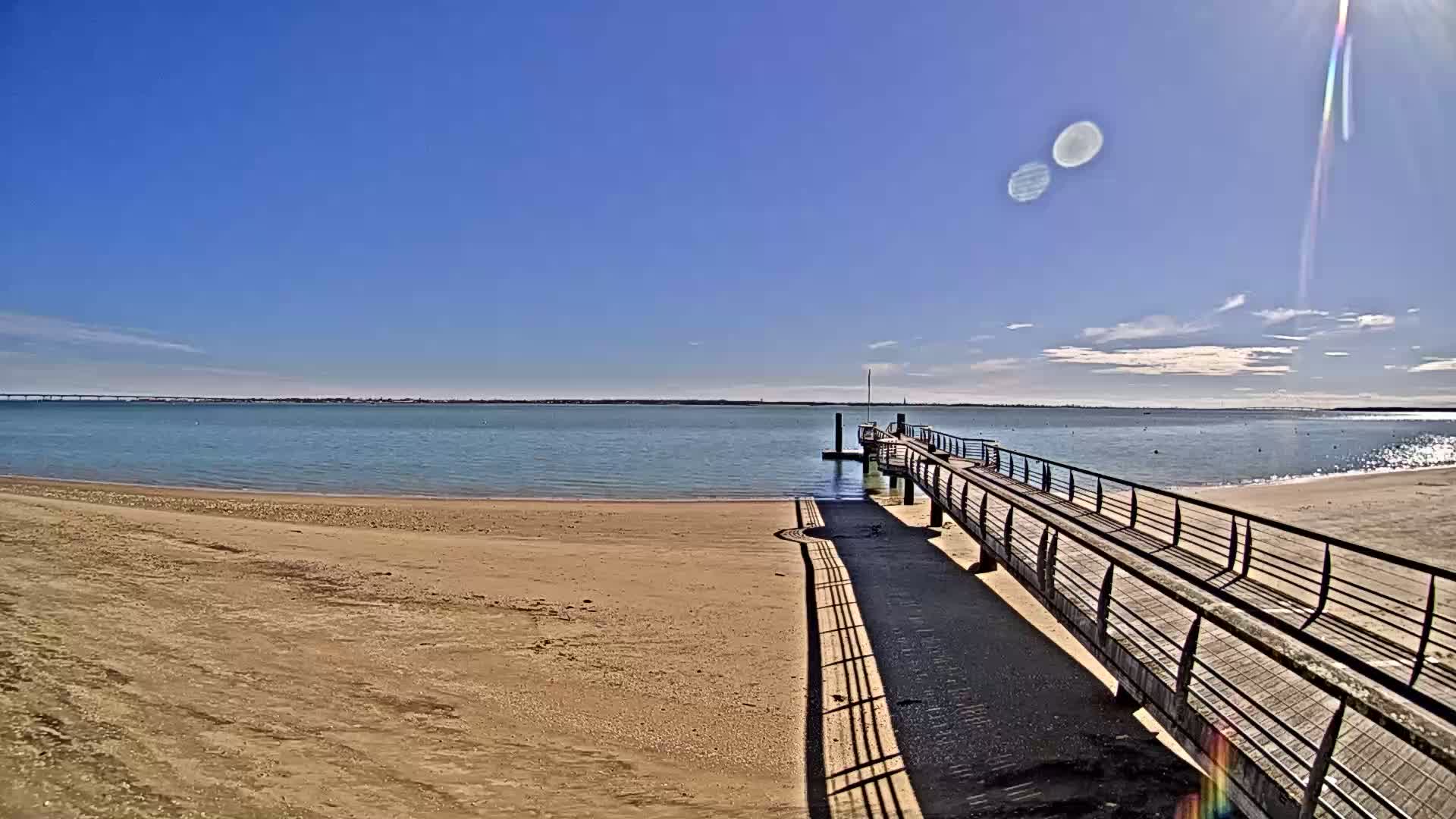 A long wooden pier extends from a sandy beach into calm, blue water under a mostly sunny sky.
