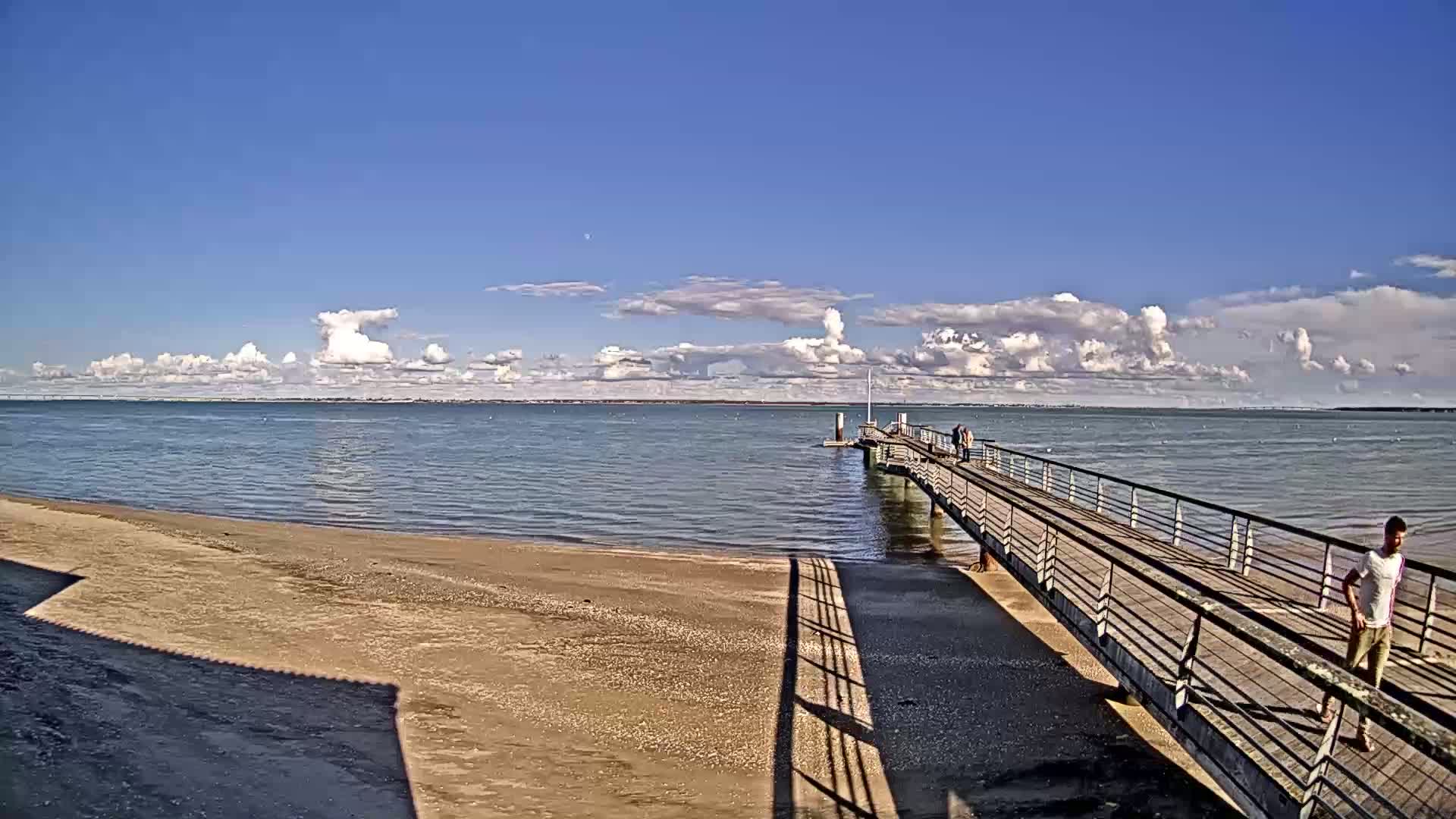 A long pier extends from a sandy beach into a calm body of water under a partly cloudy sky.