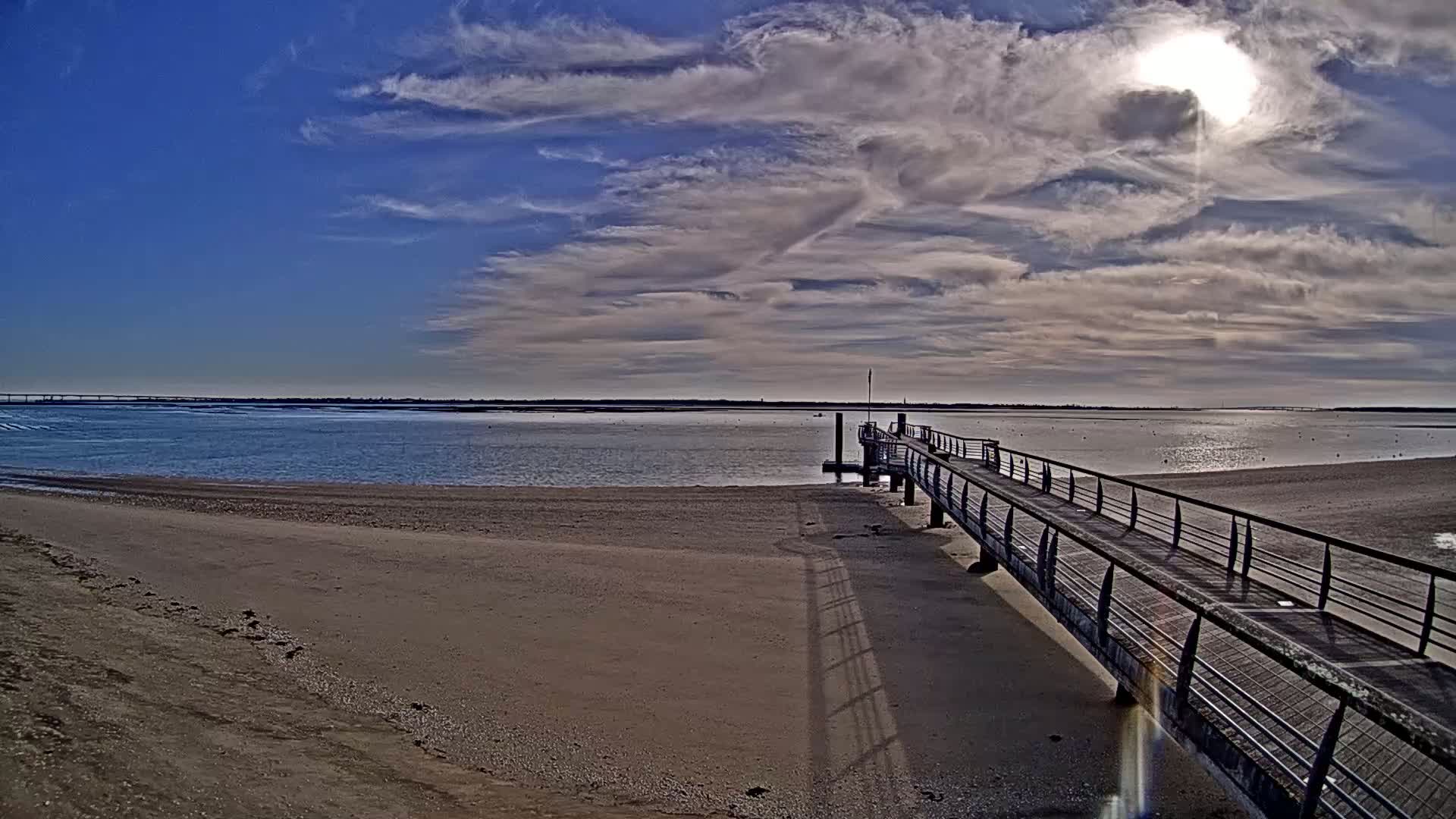 A long pier extends from a sandy beach into a calm body of water under a partly sunny sky with scattered clouds.