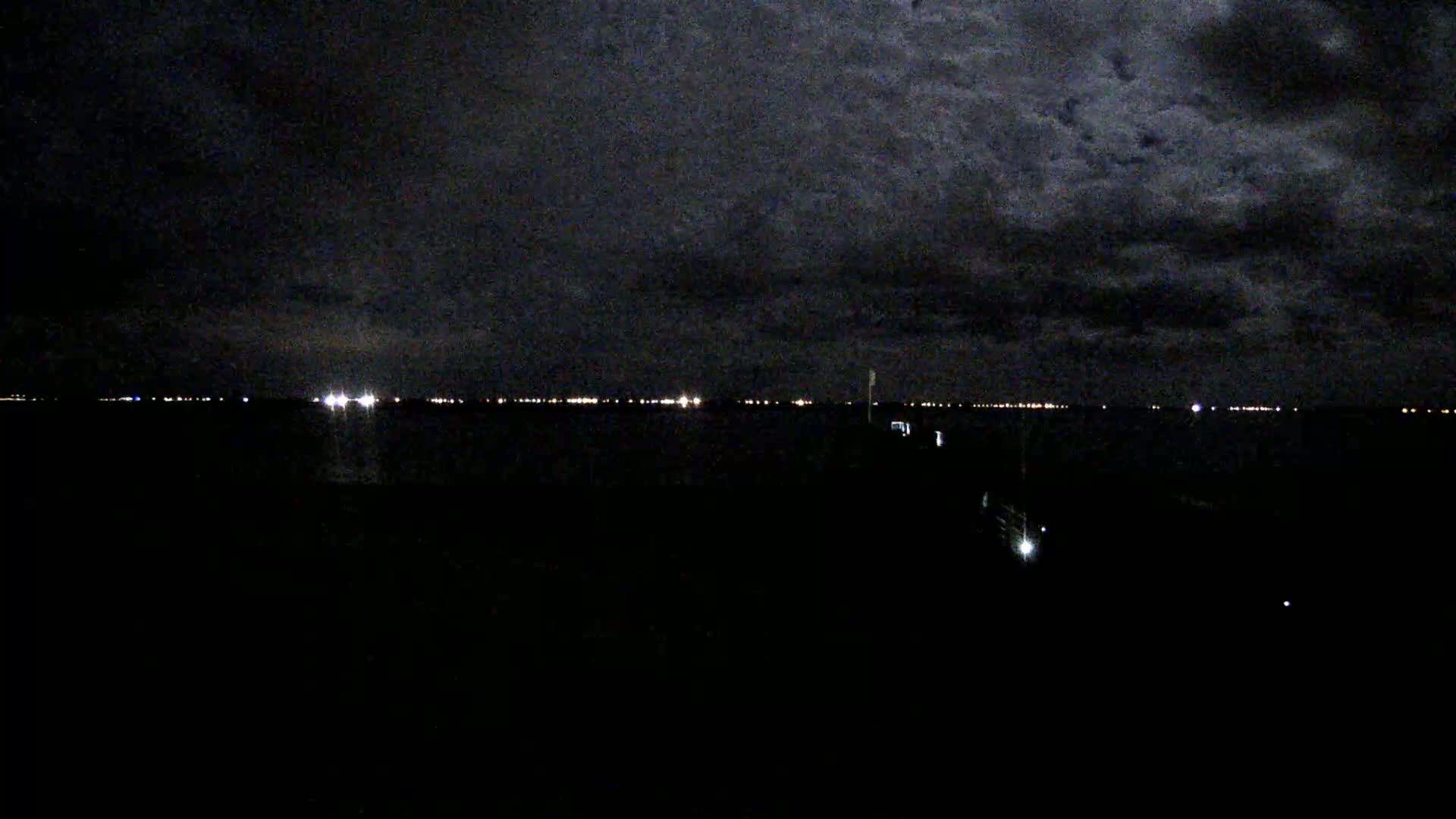 A dark, cloudy night sky overlooks a body of water with a distant line of lights on the opposite shore.