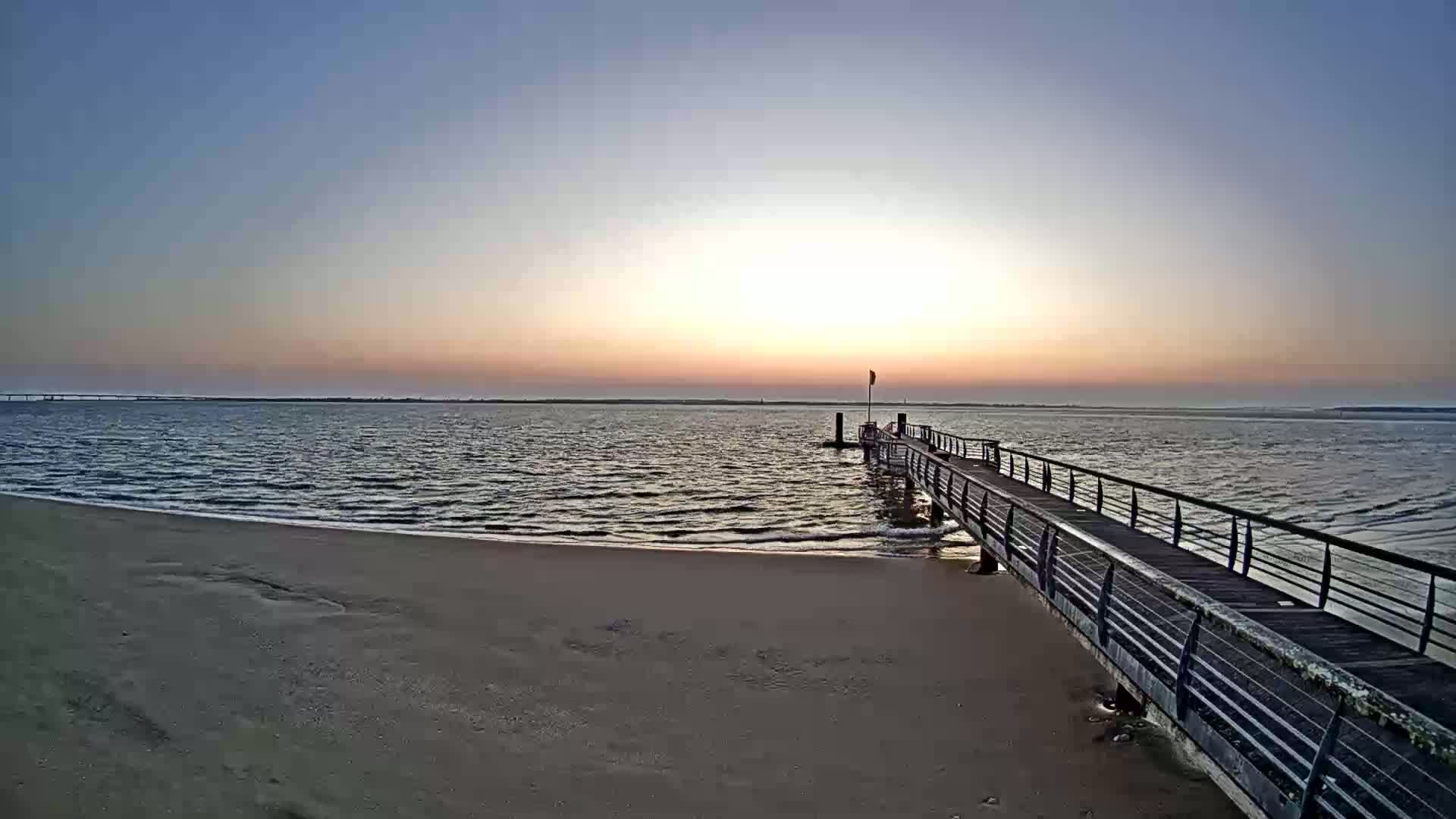 A long pier extends from a sandy beach into calm water at sunset under a clear sky.