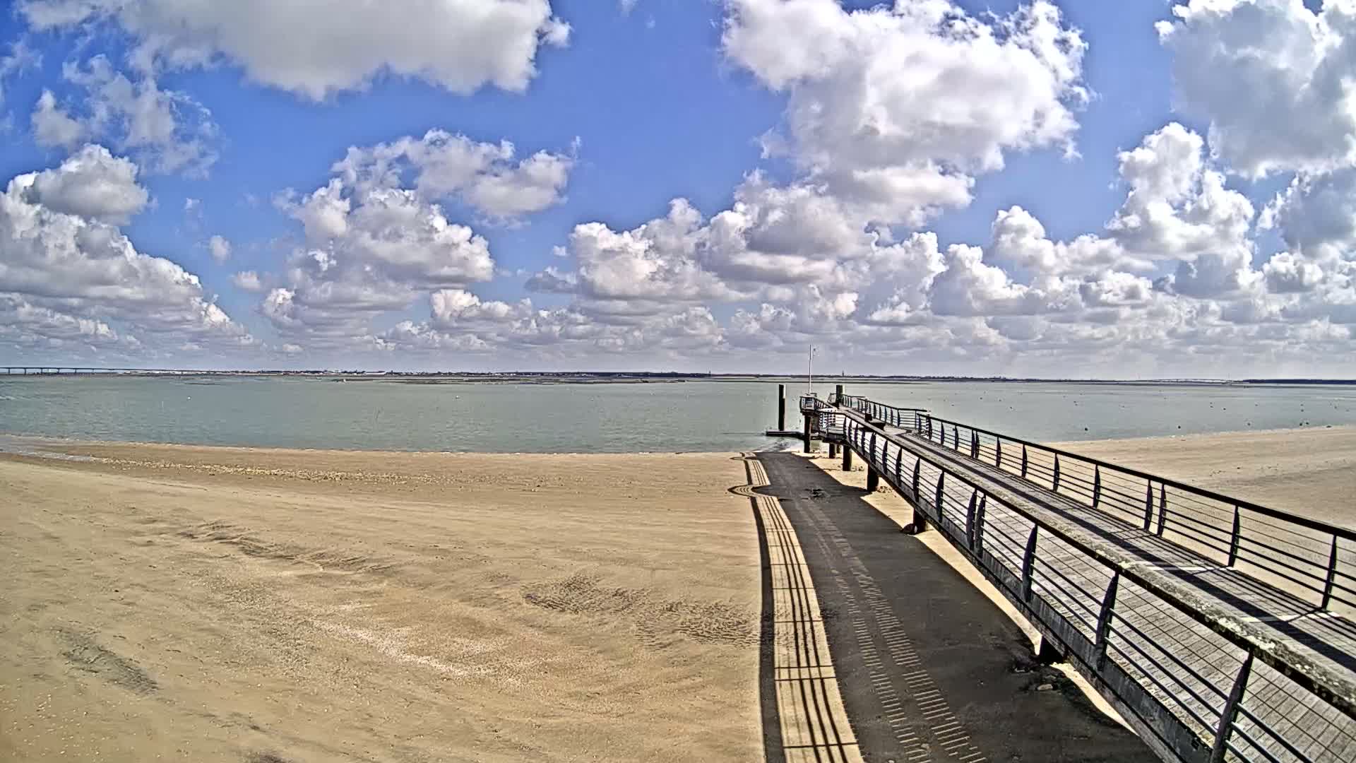 A long wooden pier extends from a sandy beach toward calm, light-green water under a partly cloudy blue sky.
