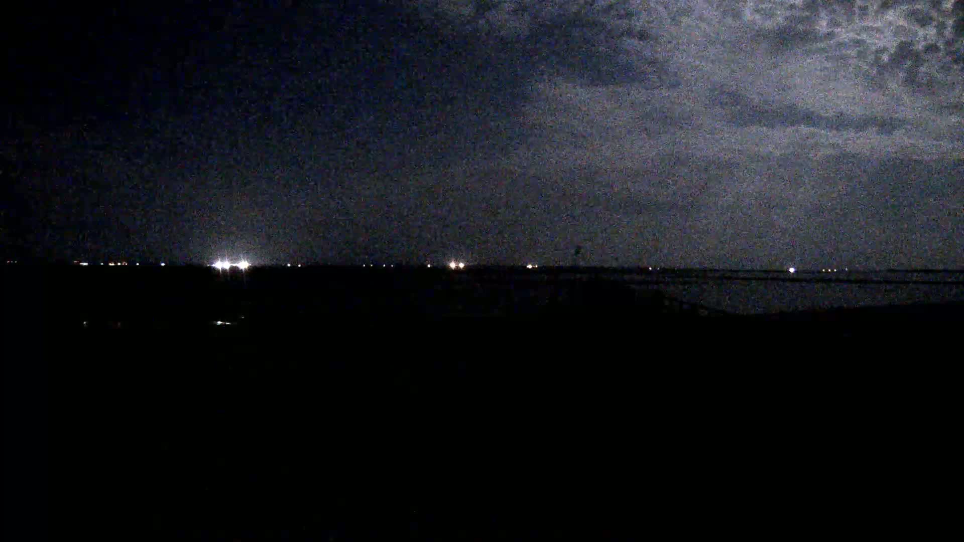 A nighttime view of a dark landscape with a distant horizon line showing faint lights under a cloudy sky.