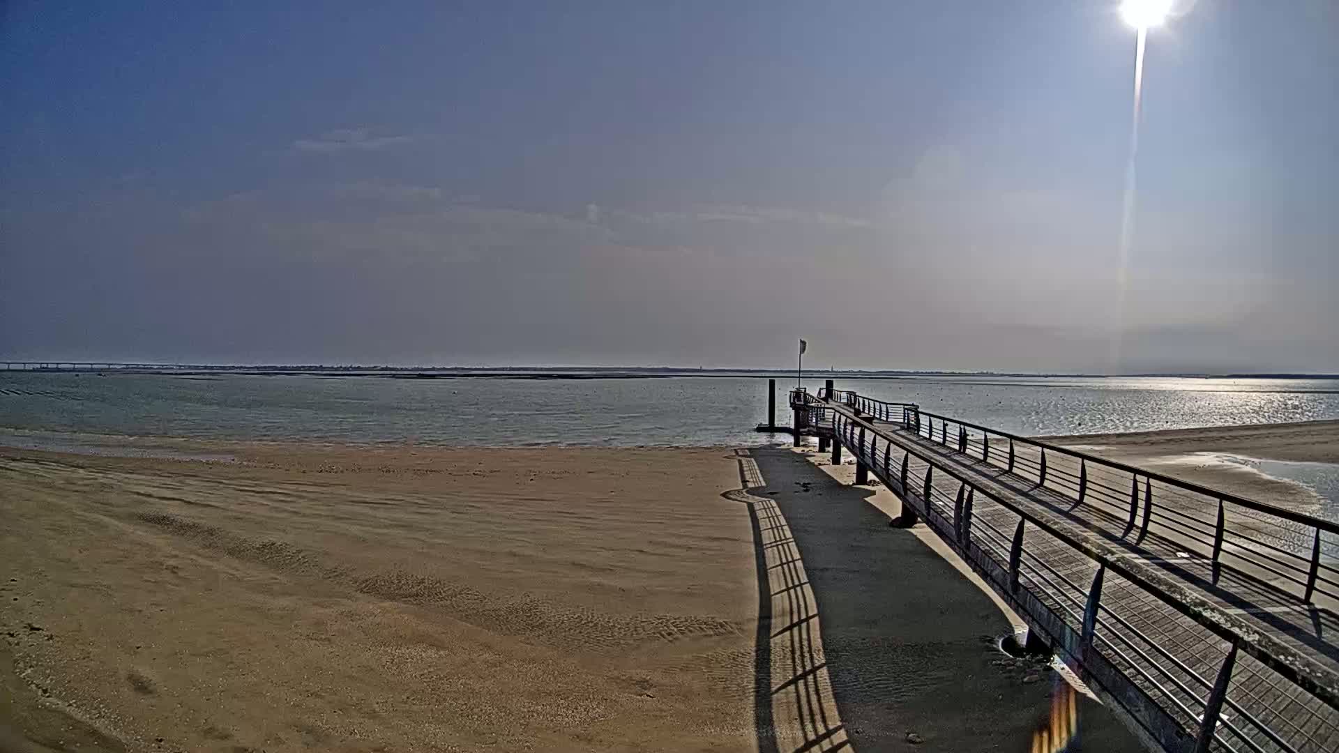 A long pier extends from a sandy beach into a calm body of water under a bright, sunny sky.