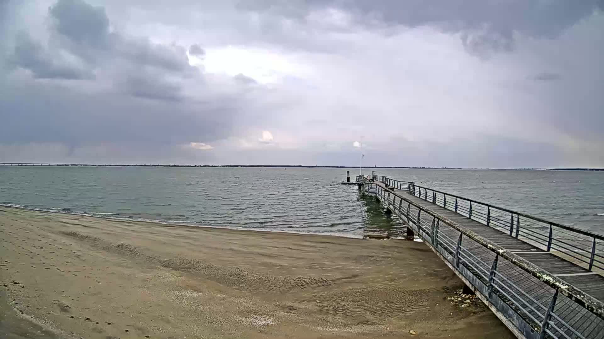 A long pier extends from a sandy beach into a calm, grey sea under an overcast sky.