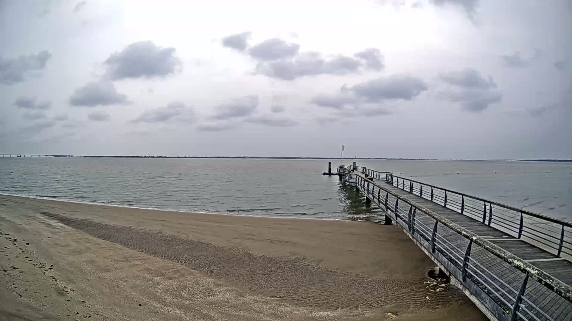 A long pier extends from a sandy beach into a calm, gray body of water under a cloudy sky.