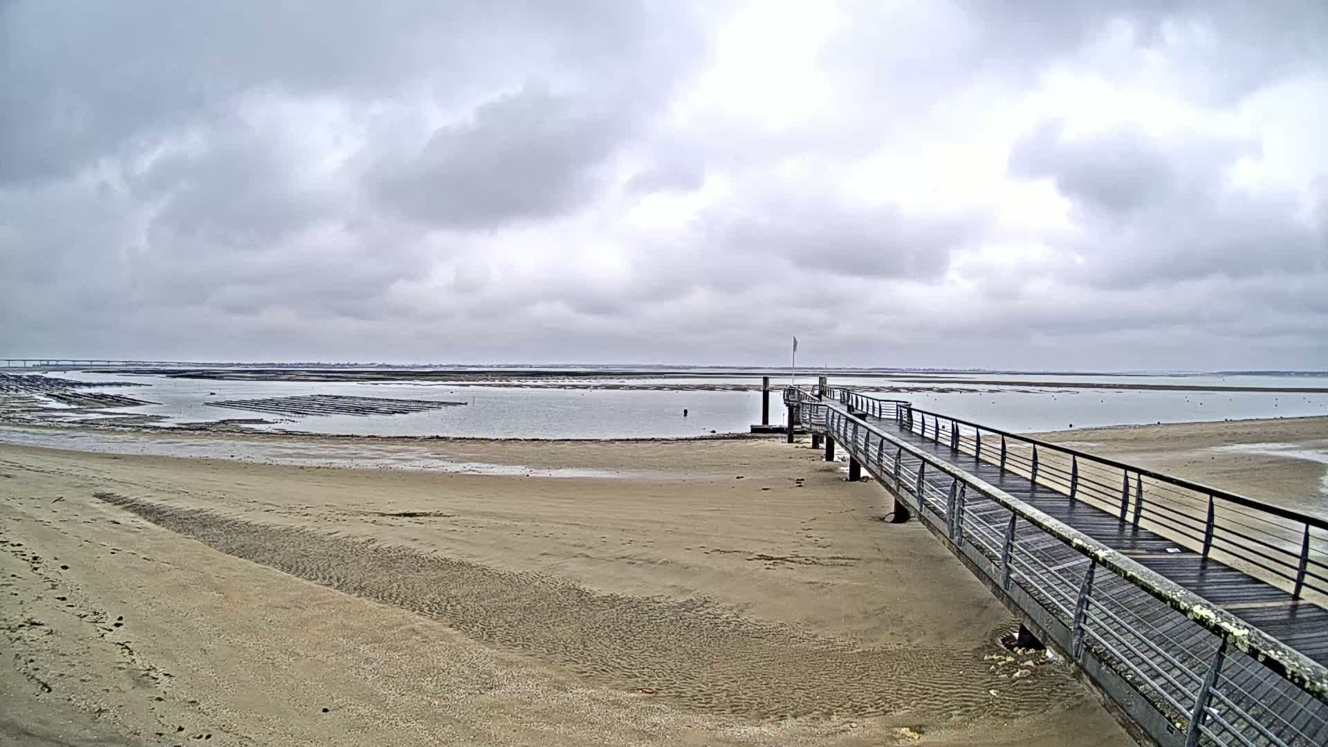 A long wooden pier extends from a sandy beach into calm, shallow water under a cloudy sky.