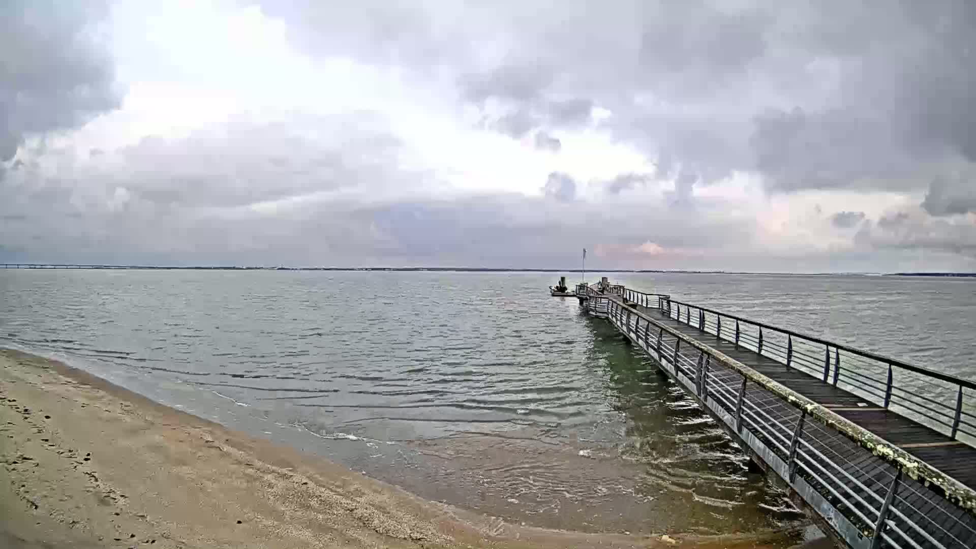 A long pier extends from a sandy beach into a choppy, gray body of water under a cloudy sky.