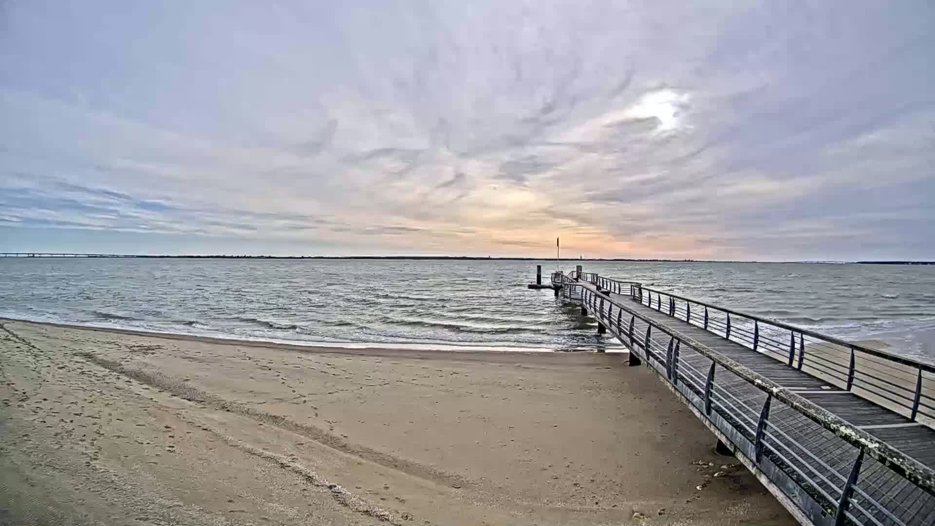 A long pier extends from a sandy beach into a choppy, grey-blue sea under a mostly cloudy sky.