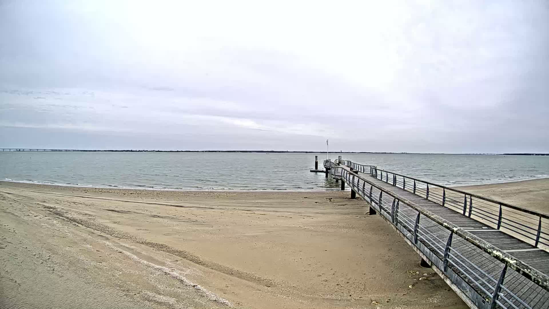 A long pier extends from a sandy beach into a calm, gray-sky body of water.