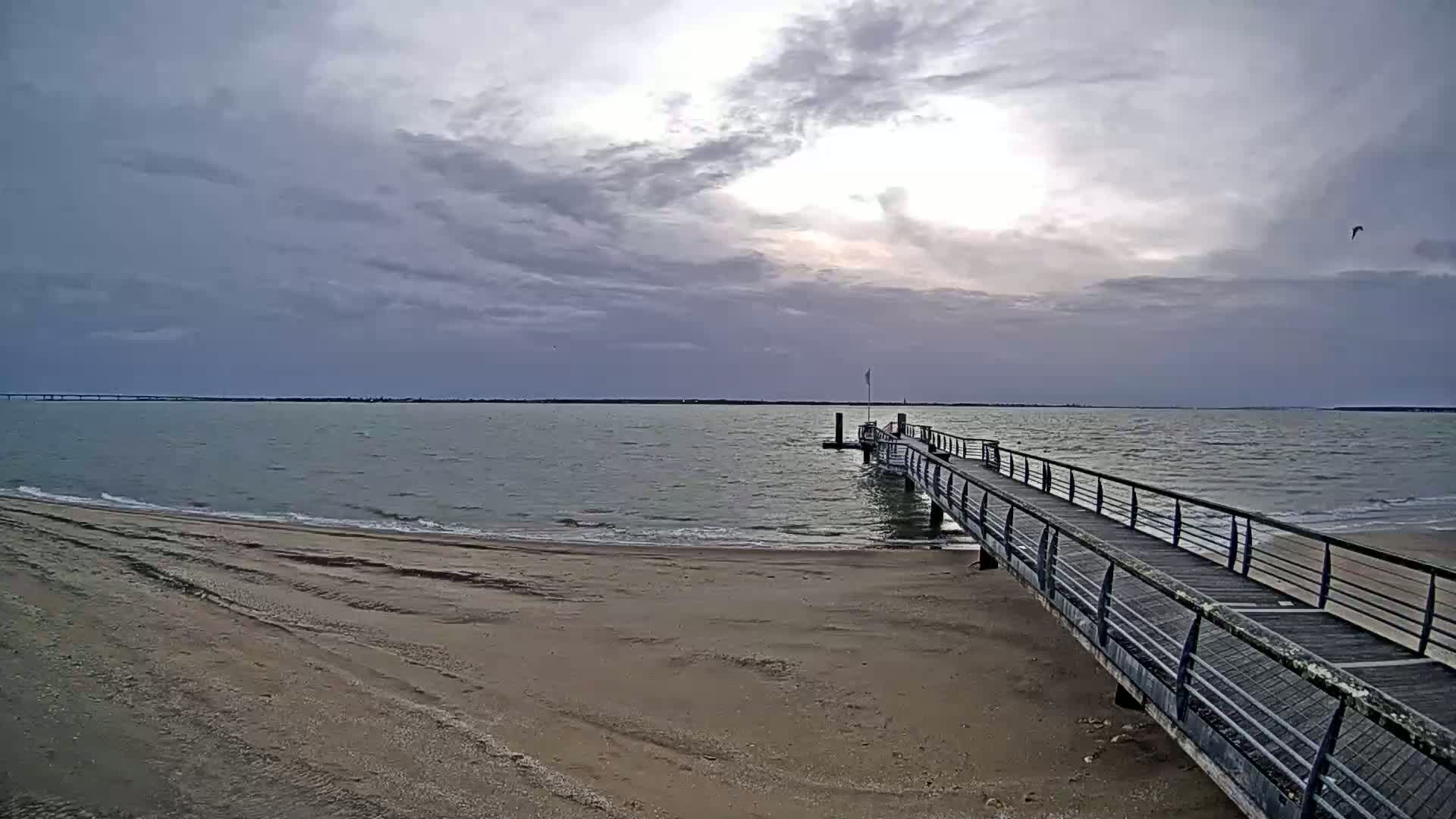 A long pier extends from a sandy beach into a calm body of water under a cloudy sky.