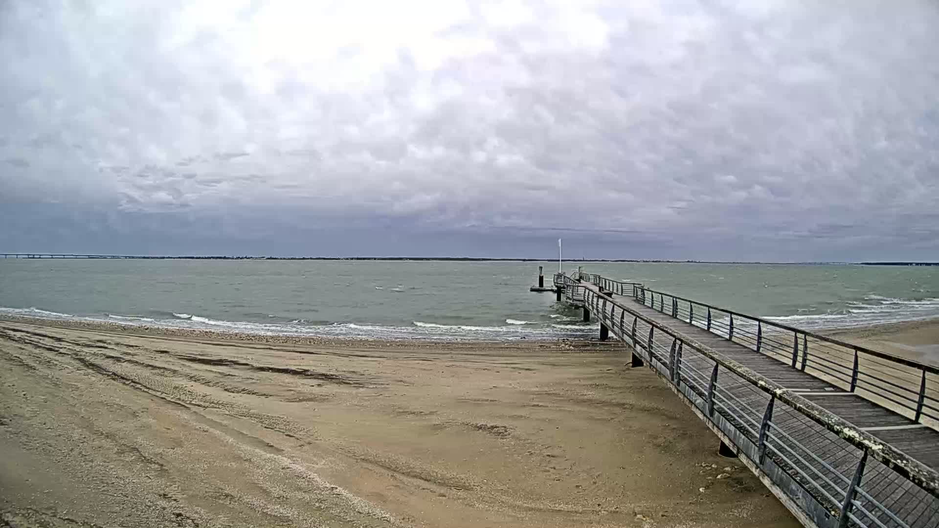 A long pier extends from a sandy beach into a choppy sea under a cloudy sky.
