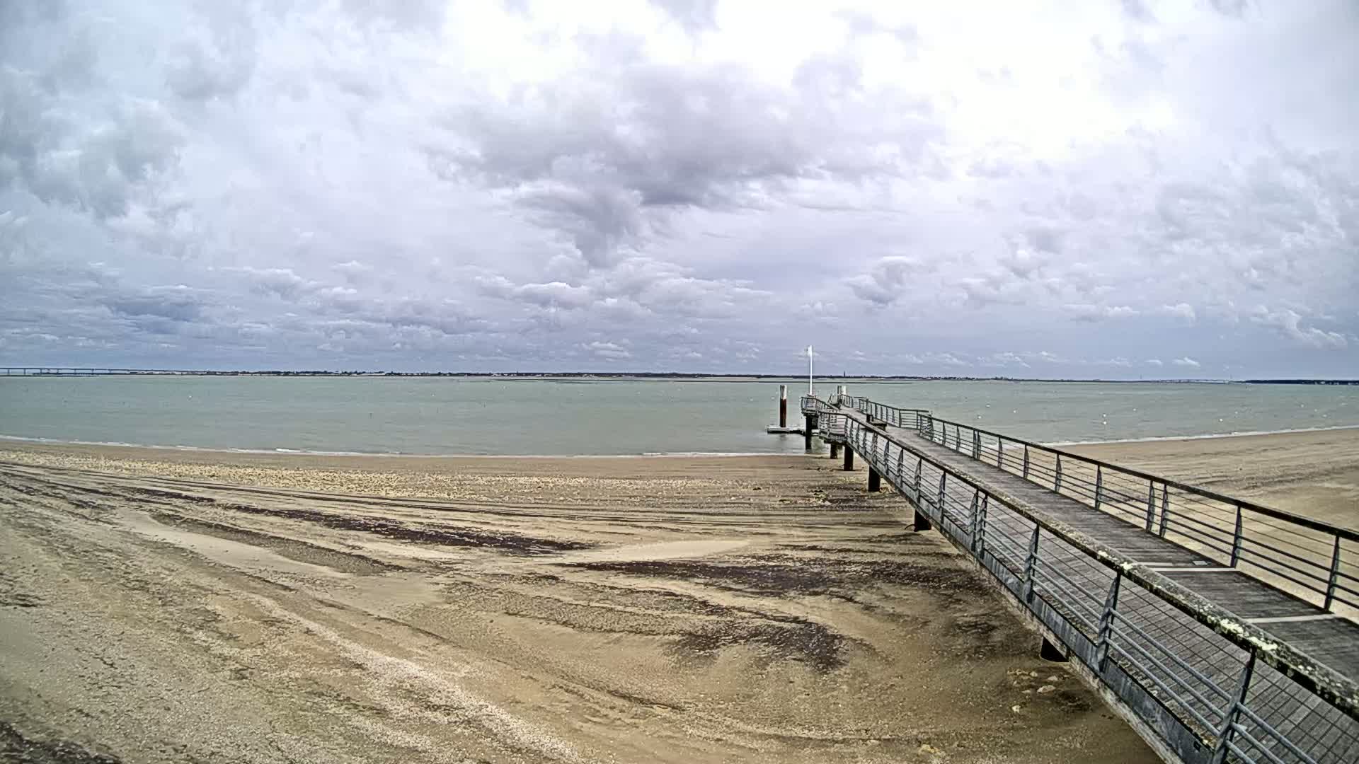 A long wooden pier extends from a sandy beach into a calm, grey-skied body of water.