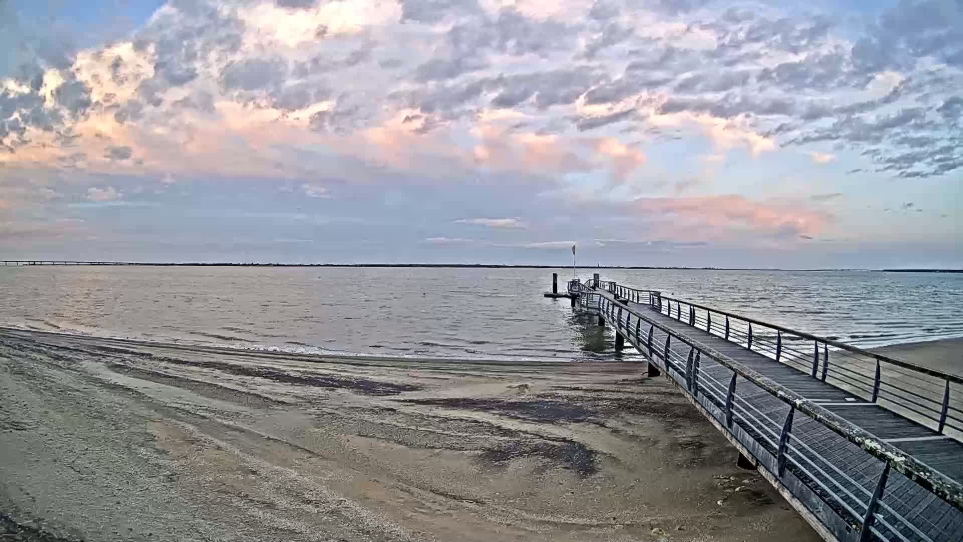 A long pier extends from a sandy beach into a calm body of water under a partly cloudy sky with pink hues near the horizon.