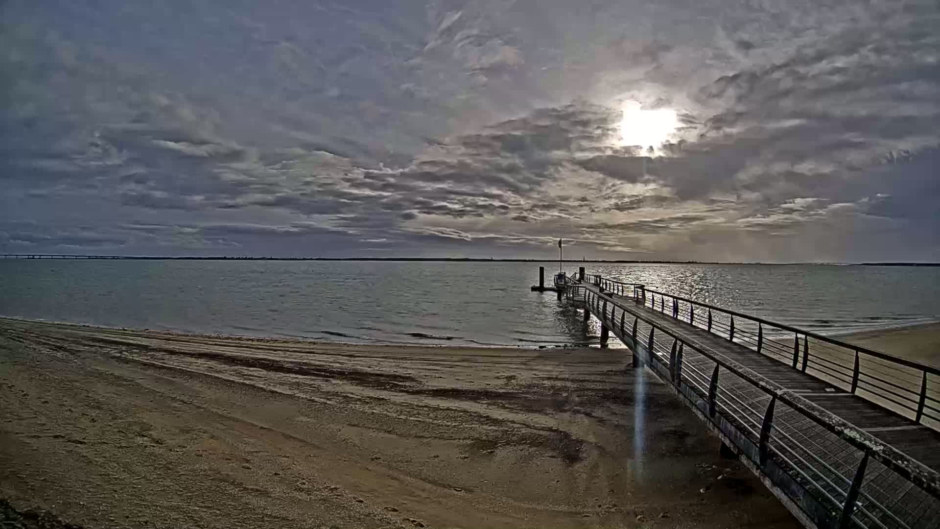 A long pier extends from a sandy beach into a calm body of water under a partly cloudy sky, with the sun shining through the clouds.