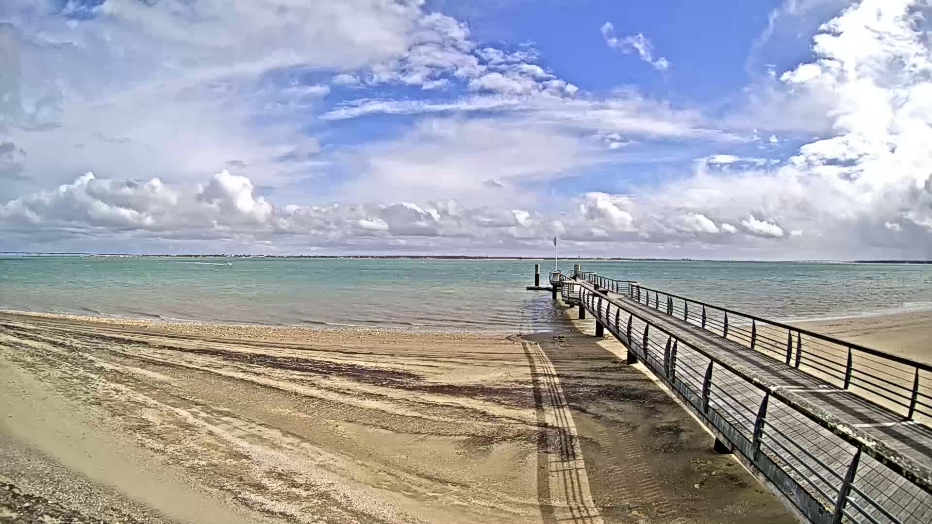 A long pier extends from a sandy beach into a calm body of water under a partly cloudy sky.