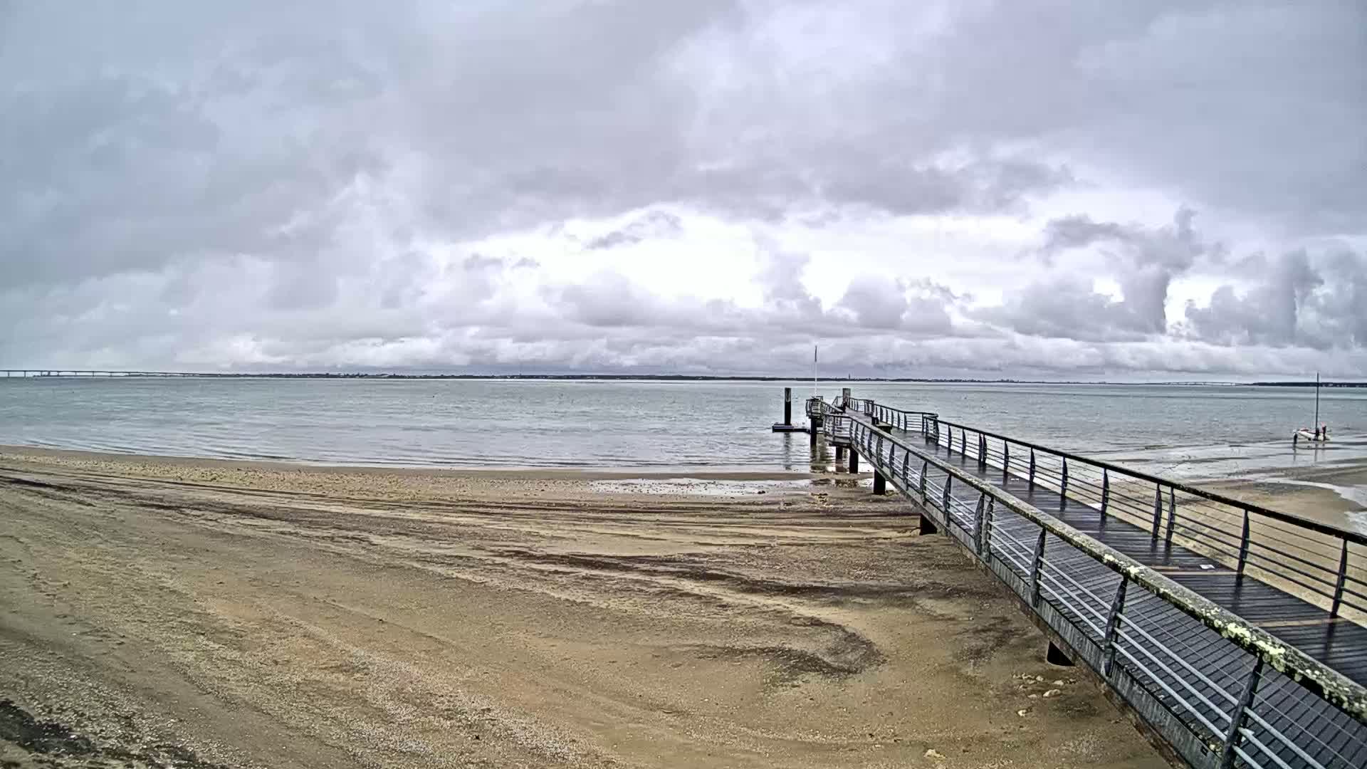 A sandy beach stretches toward a long pier extending into a calm body of water under a cloudy sky.