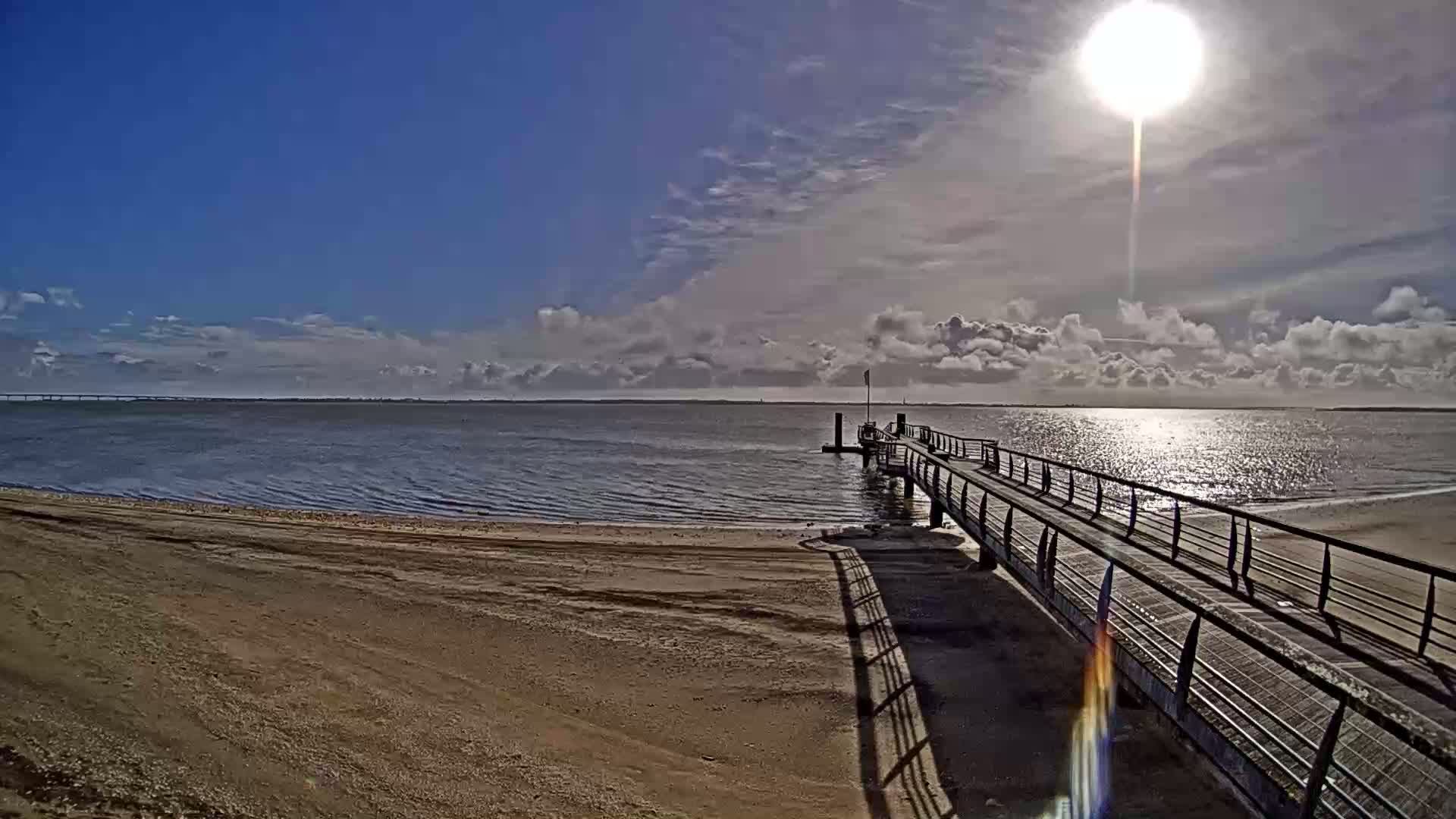 A sandy beach leads to a long pier extending into a calm body of water under a partly cloudy, sunny sky.