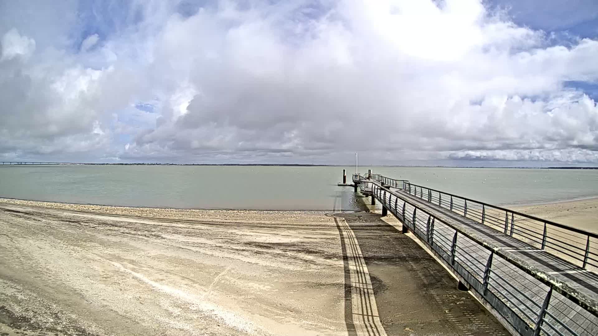 A long pier extends from a sandy beach into a calm body of water under a partly cloudy sky.