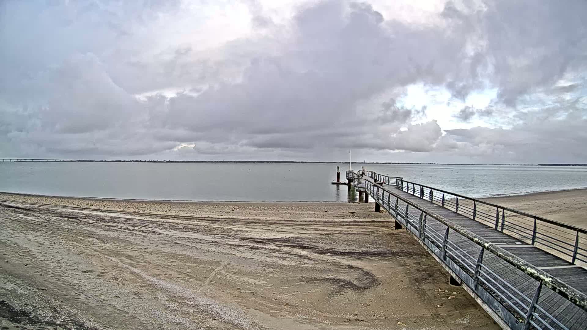 A long pier extends from a sandy beach into a calm body of water under a cloudy sky.