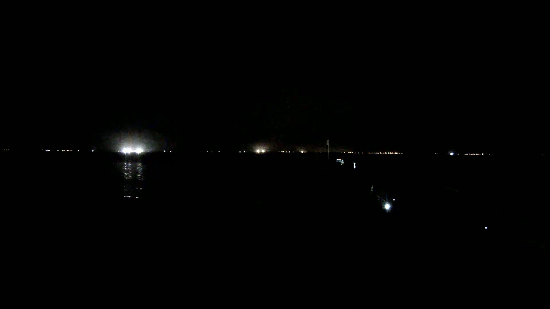 A dark, nighttime scene shows a body of water reflecting distant lights on a horizon line.