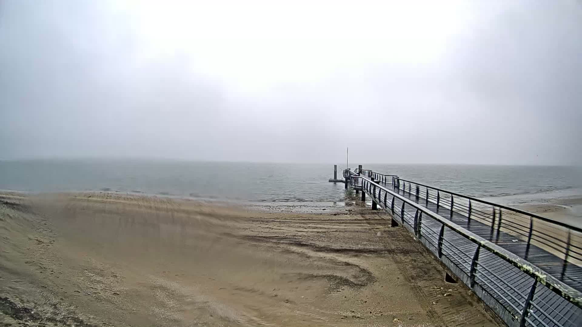 A long pier extends from a sandy beach into a foggy, gray sea.