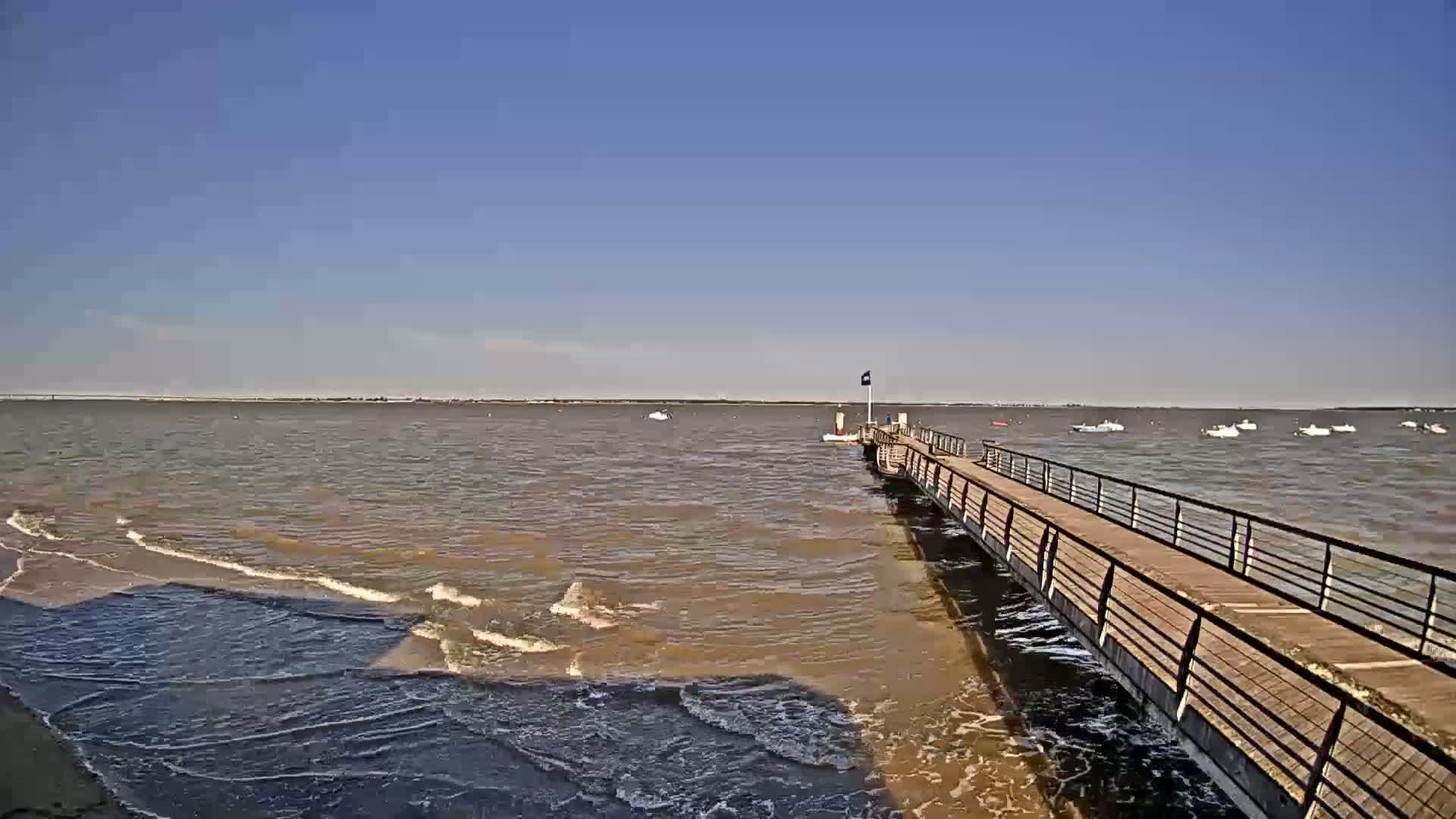 A long pier extends into a calm, brown-tinged body of water under a clear blue sky, with several small boats visible in the distance.
